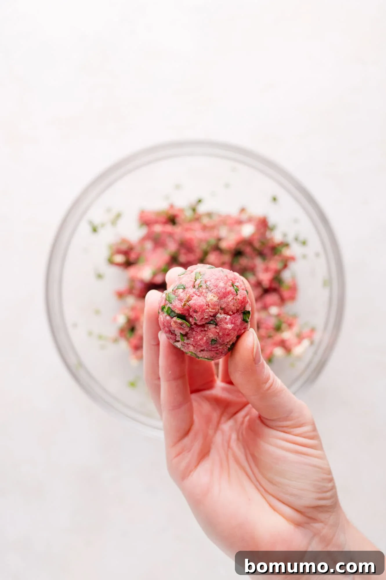 A hand holds a raw meatball in front of a glass bowl filled with ground meat mixture.