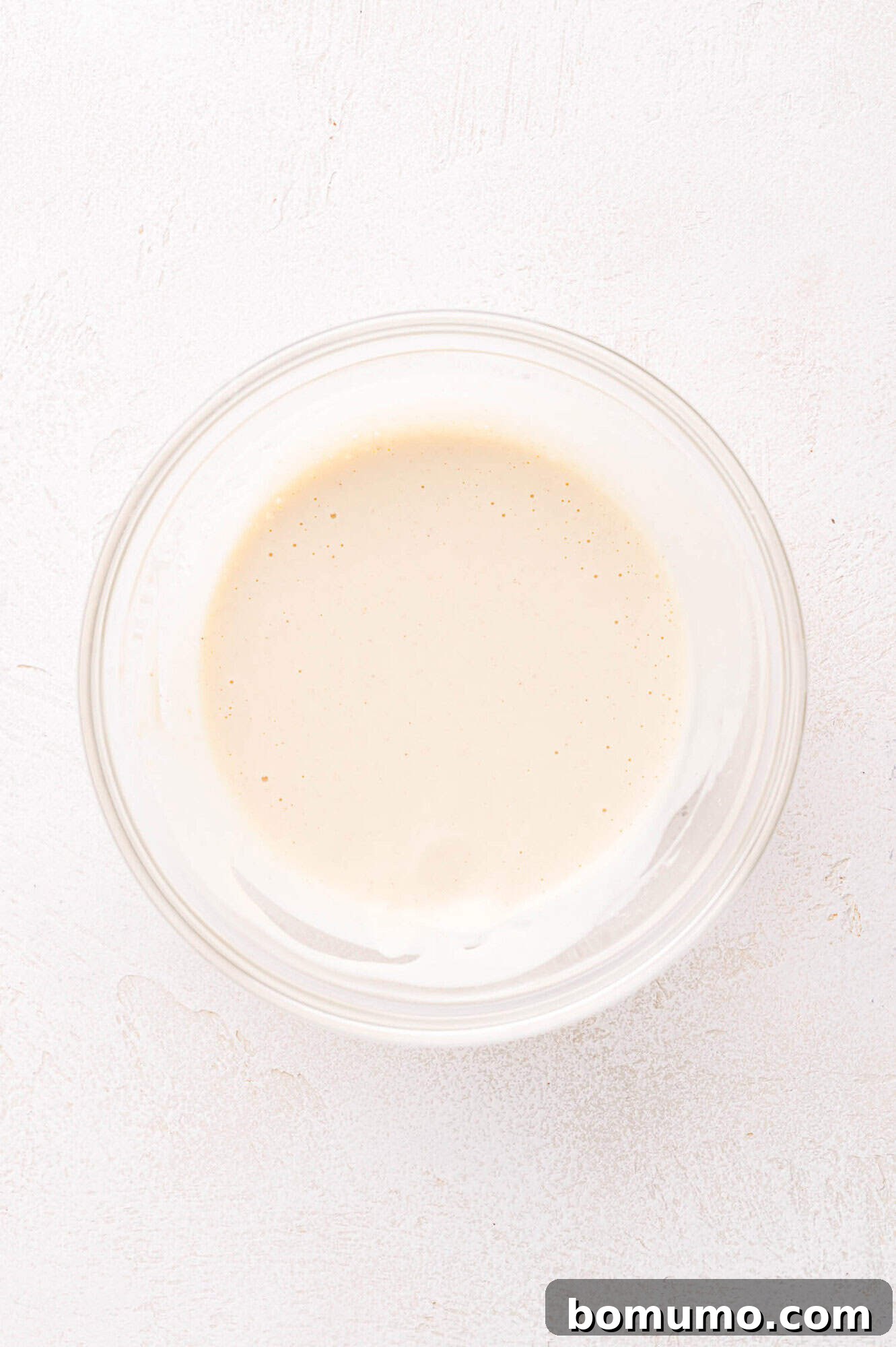 A small, clear glass bowl filled with a smoothly blended, creamy, light beige sauce, resting on a textured white surface, ready to be spread.