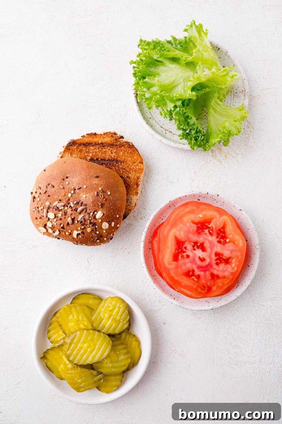 A pristine white plate holds a vibrant array of fresh toppings: crisp green lettuce, a perfectly sliced wheat burger bun, a bowl brimming with tangy pickle slices, and a separate plate showcasing two bright red tomato slices, all arranged artfully on a white surface.