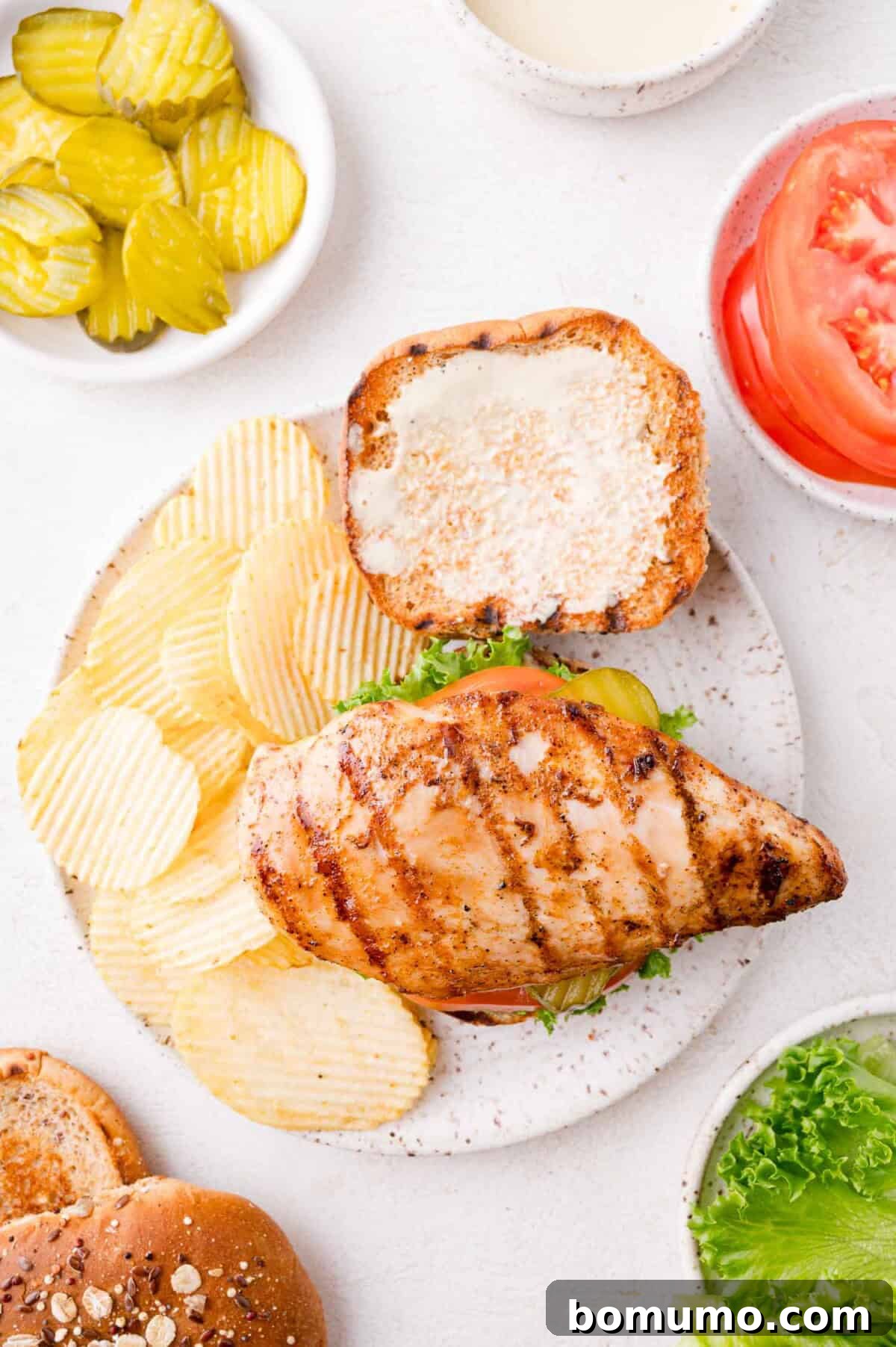 A beautifully assembled grilled chicken sandwich, topped with fresh lettuce on a toasted bun, served alongside perfectly ridged potato chips, vibrant sliced pickles, and ripe tomato slices, with a small bowl of our signature sauce for dipping, all arranged on a clean white surface.