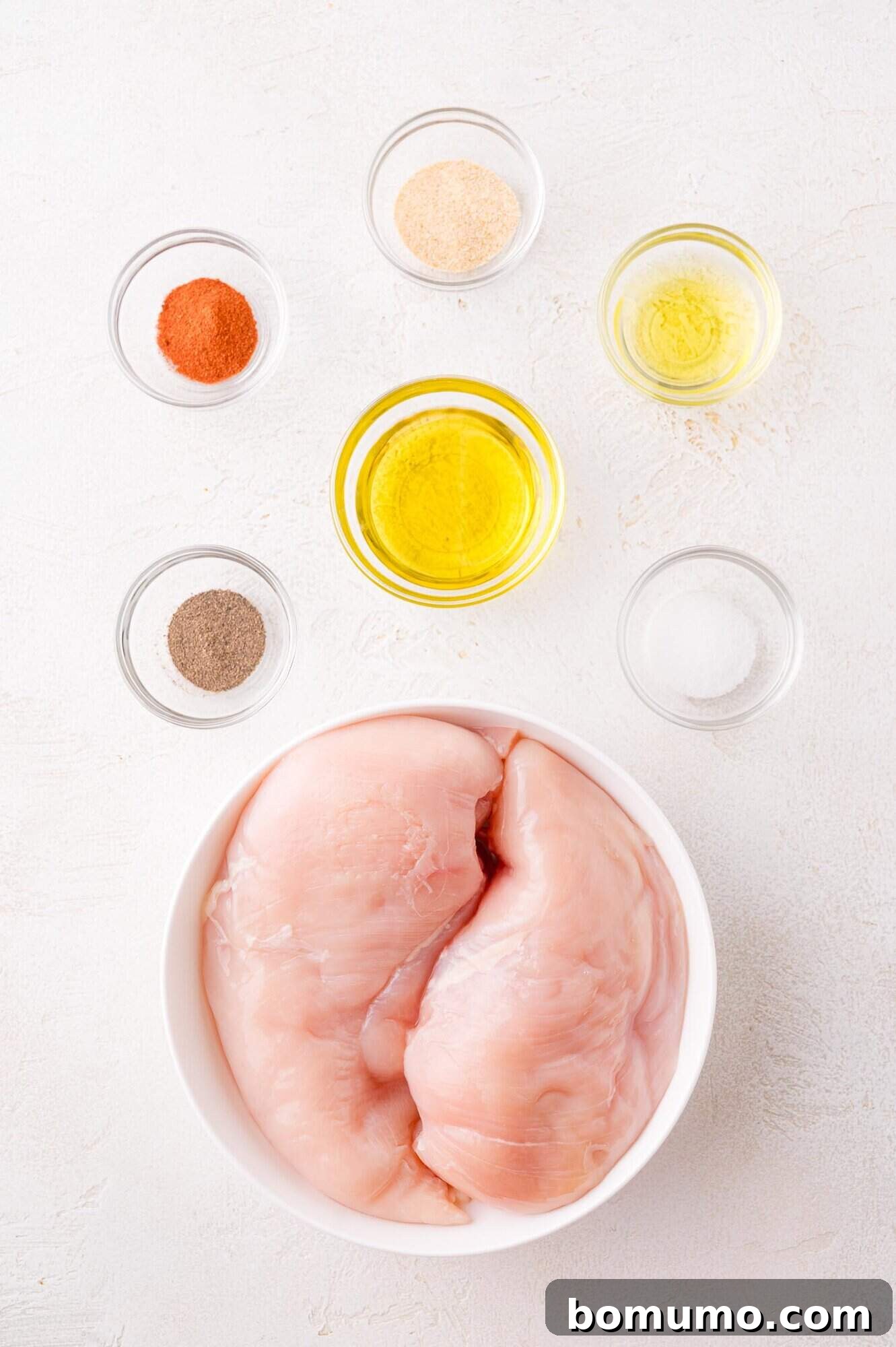 Raw chicken breasts in a white bowl, surrounded by small glass bowls containing various spices, olive oil, and seasoning, all arranged on a pristine white surface.