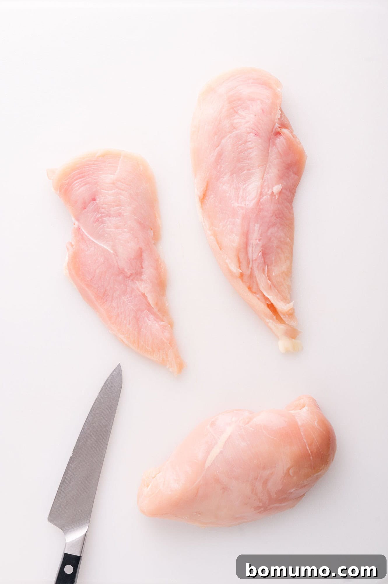 A raw whole chicken breast beside two perfectly sliced chicken breast halves, displayed on a white cutting board with a kitchen knife nearby, illustrating the preparation step.