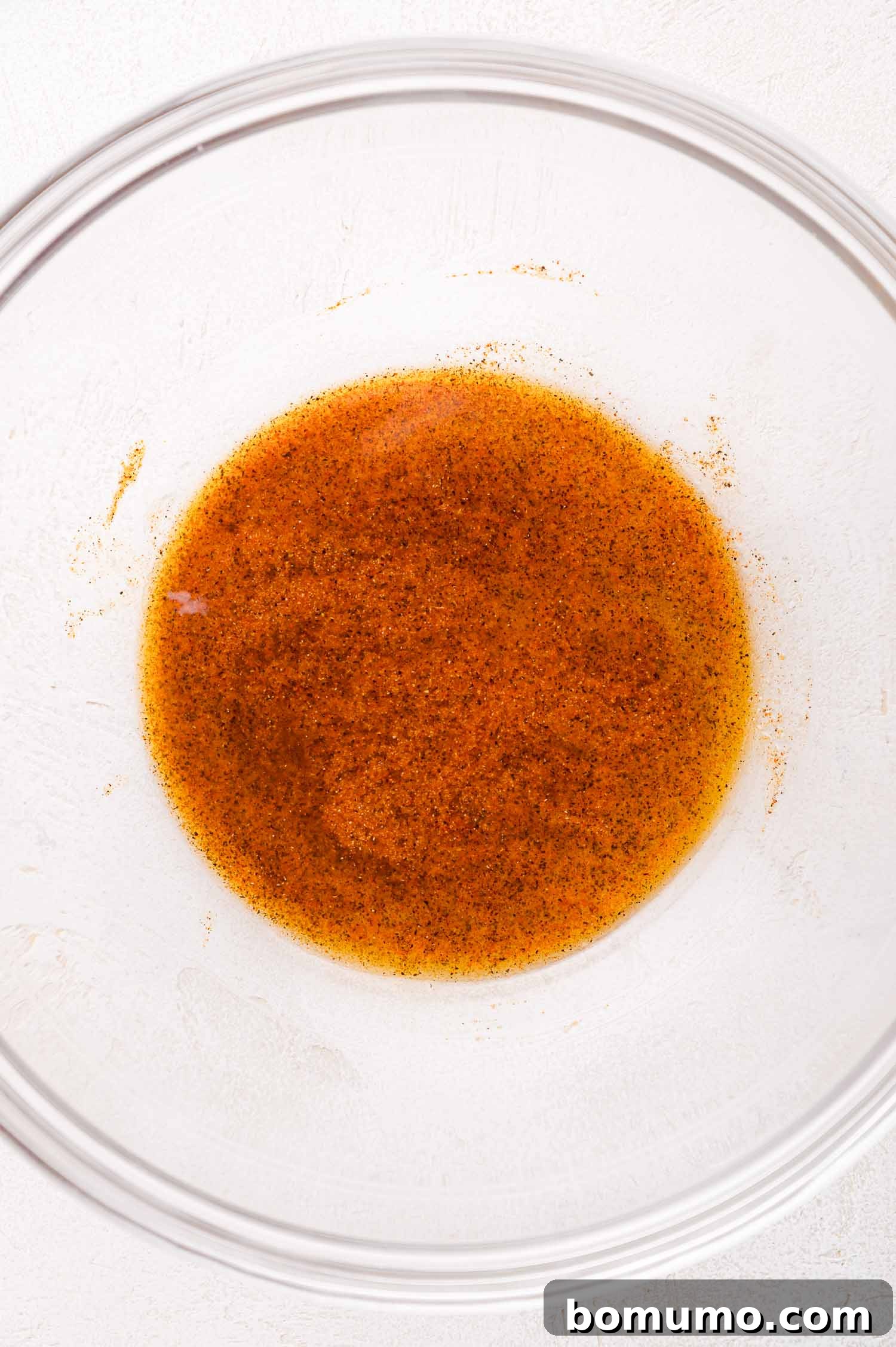 A clear glass bowl showcases a rich brown marinade, flecked with spices, resting on a clean white surface, ready for the chicken.