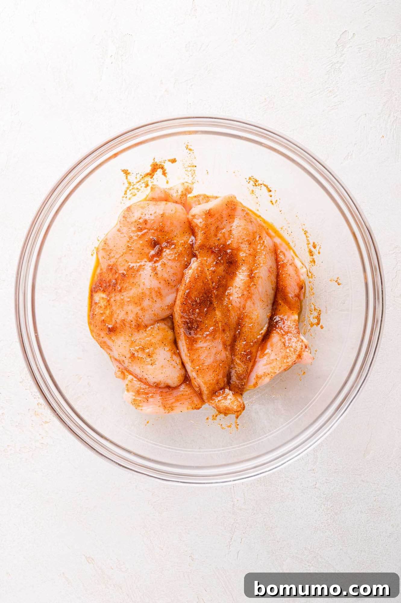 Two raw chicken breasts, glistening with a savory marinade and seasoning, presented in a clear glass bowl on a light-colored surface, awaiting their turn on the grill.