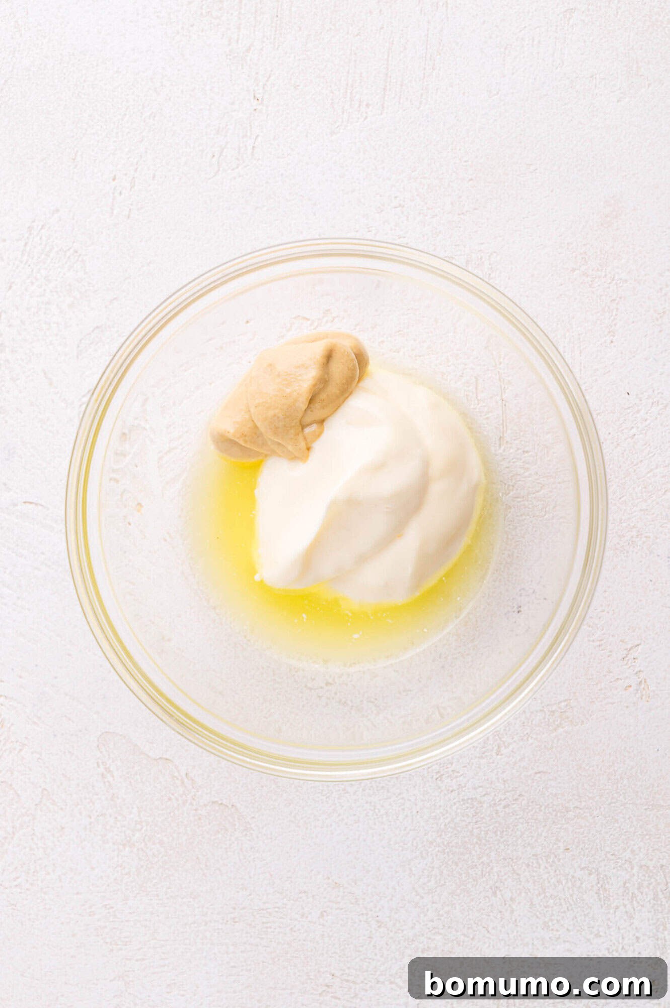 A clear glass bowl holding a mixture of creamy mayonnaise, vibrant yellow mustard, and a splash of pickle juice, poised on a light-colored surface.