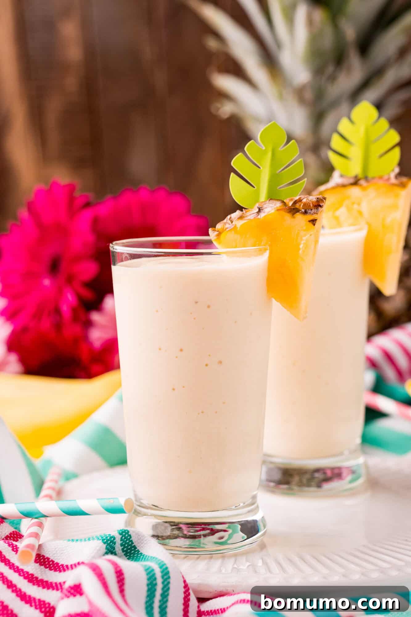 Pineapple Paradise Smoothie 2 Two glasses of creamy pineapple smoothie garnished with pineapple wedges and green leaf stirrers, with a colorful striped cloth and pink flowers in the background.