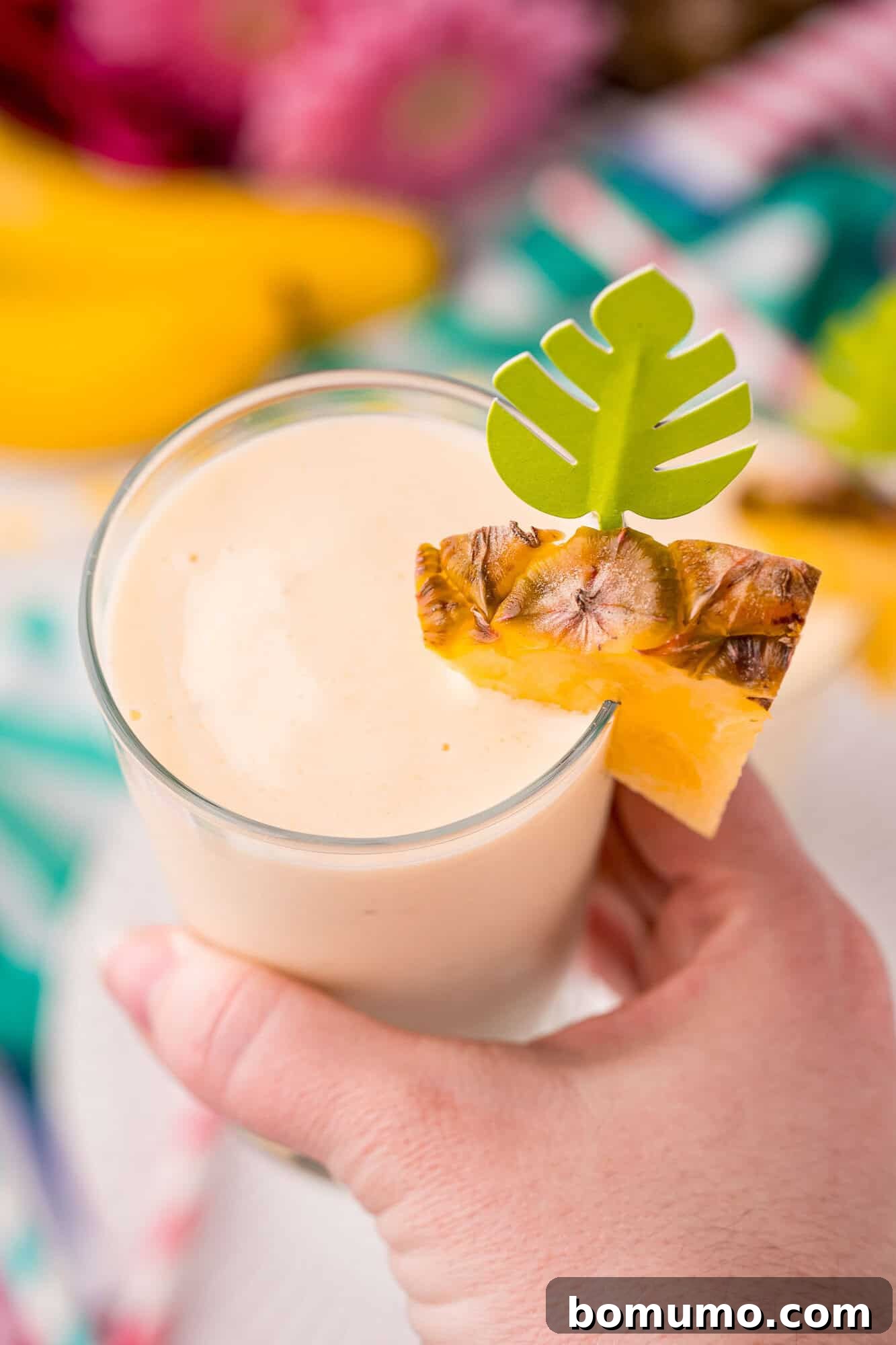Pineapple Paradise Smoothie 3 A hand holds a glass of creamy smoothie garnished with a pineapple slice and a decorative green paper leaf.