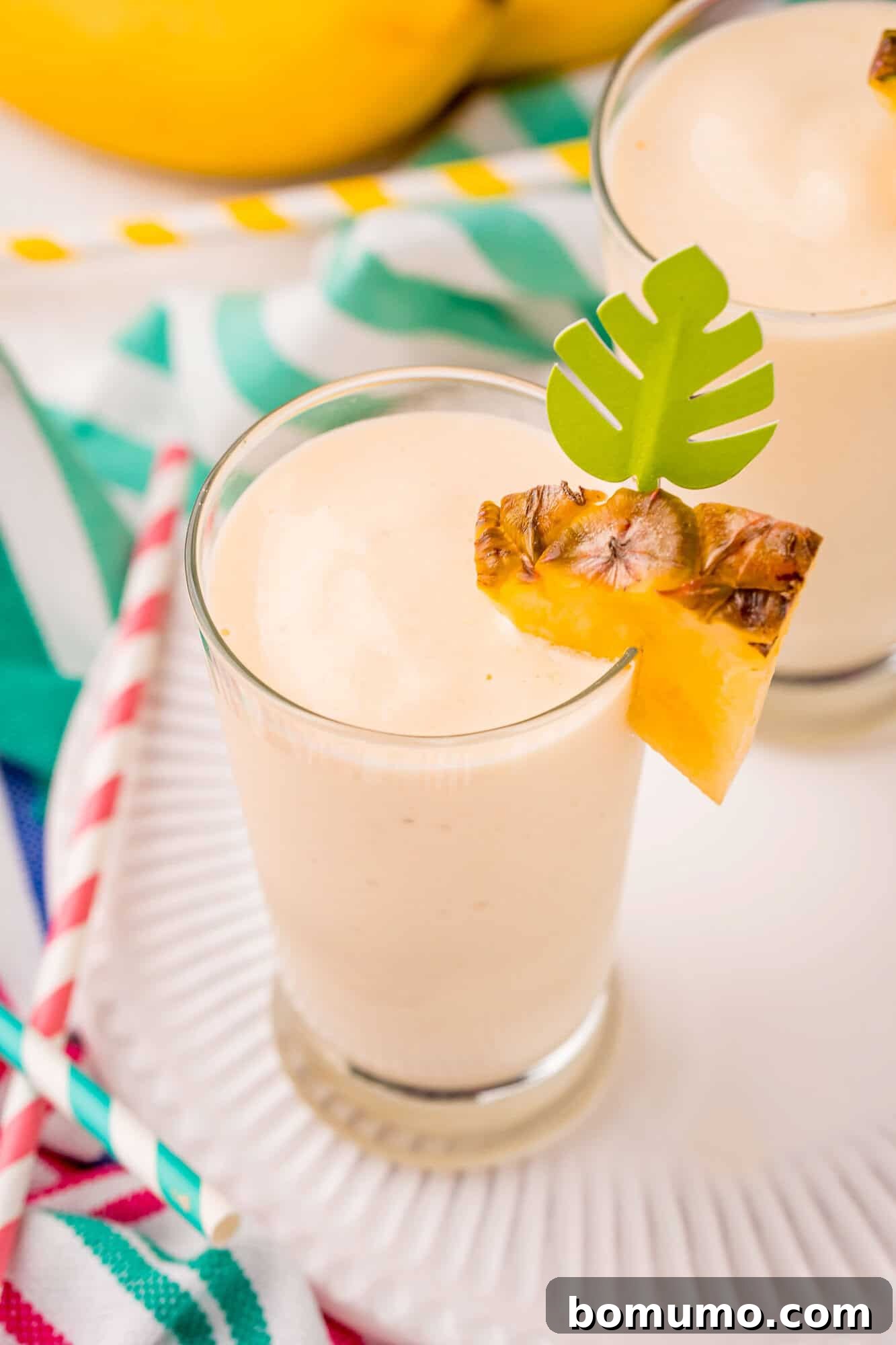 Pineapple Paradise Smoothie 5 A glass of creamy smoothie garnished with a pineapple slice and a green paper leaf, placed on a white plate with colorful straws nearby.