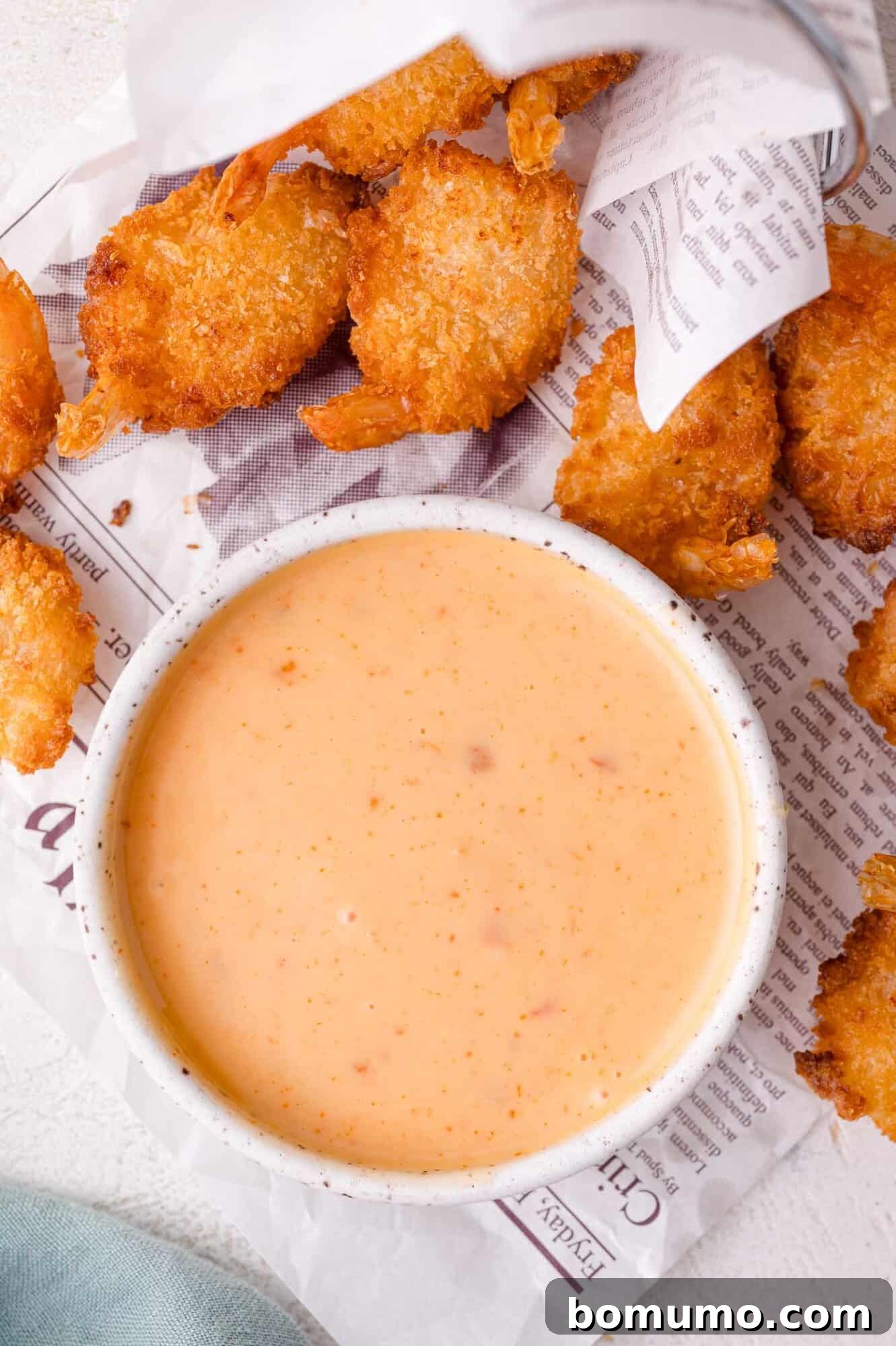 Sweet Chili Blast 2 A elegant bowl of creamy, orange-hued Bang Bang dipping sauce, artfully placed on a newspaper-style surface, surrounded by several pieces of perfectly fried and breaded shrimp, ready to be enjoyed.