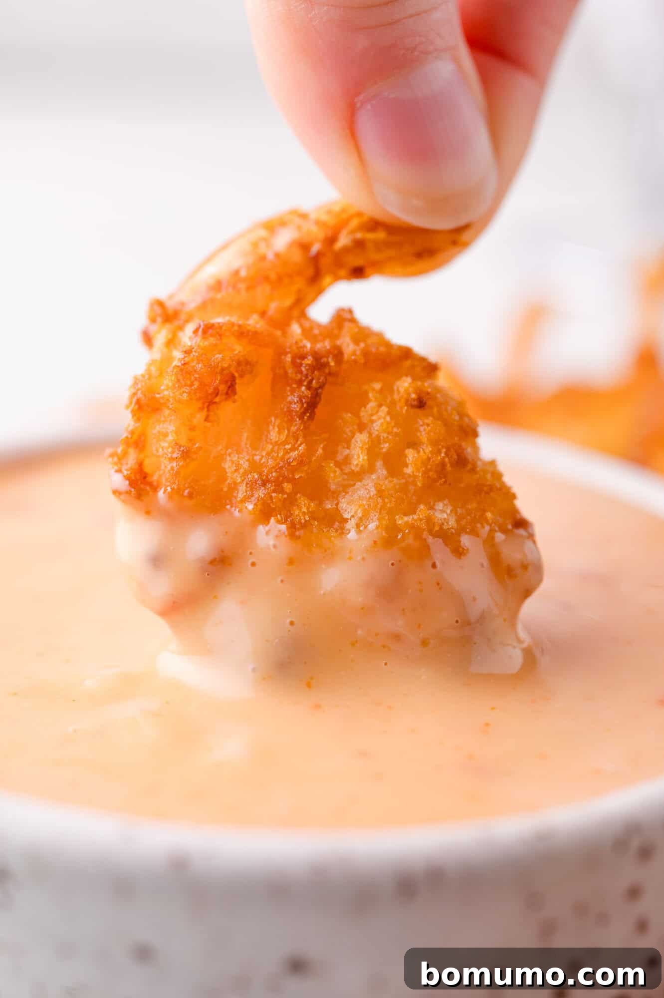 Sweet Chili Blast 3 A hand gently dips a piece of golden, crispy fried shrimp into a small, elegant white bowl brimming with creamy, vibrant orange-colored Bang Bang Sauce, showcasing its perfect dipping consistency.