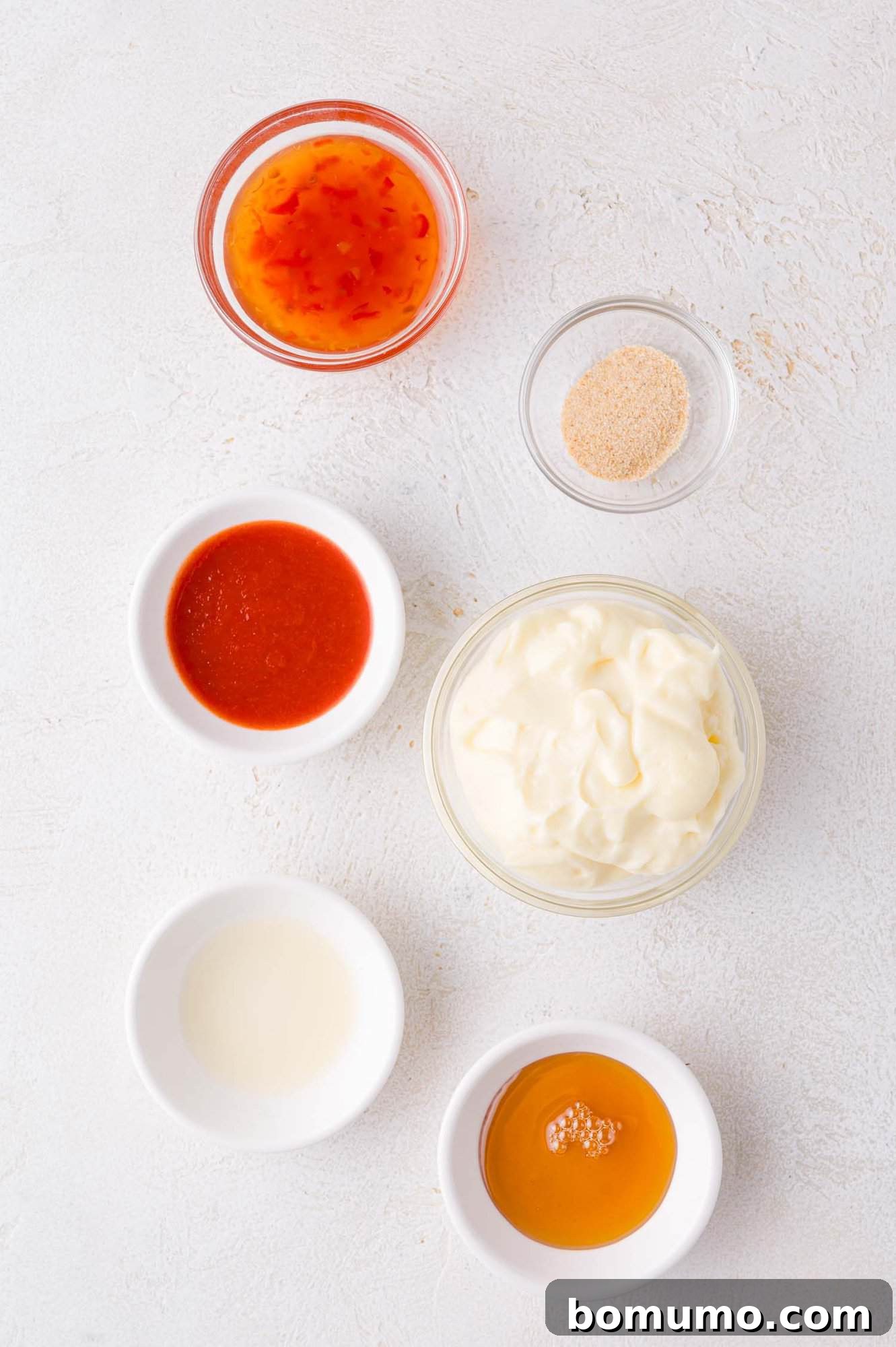 Sweet Chili Blast 4 Six small, pristine white bowls are carefully arranged on a clean white surface, each containing a distinct ingredient for Bang Bang Sauce: creamy mayonnaise, vibrant sweet chili sauce, fiery sriracha, finely ground garlic powder, clear vinegar, and golden honey.