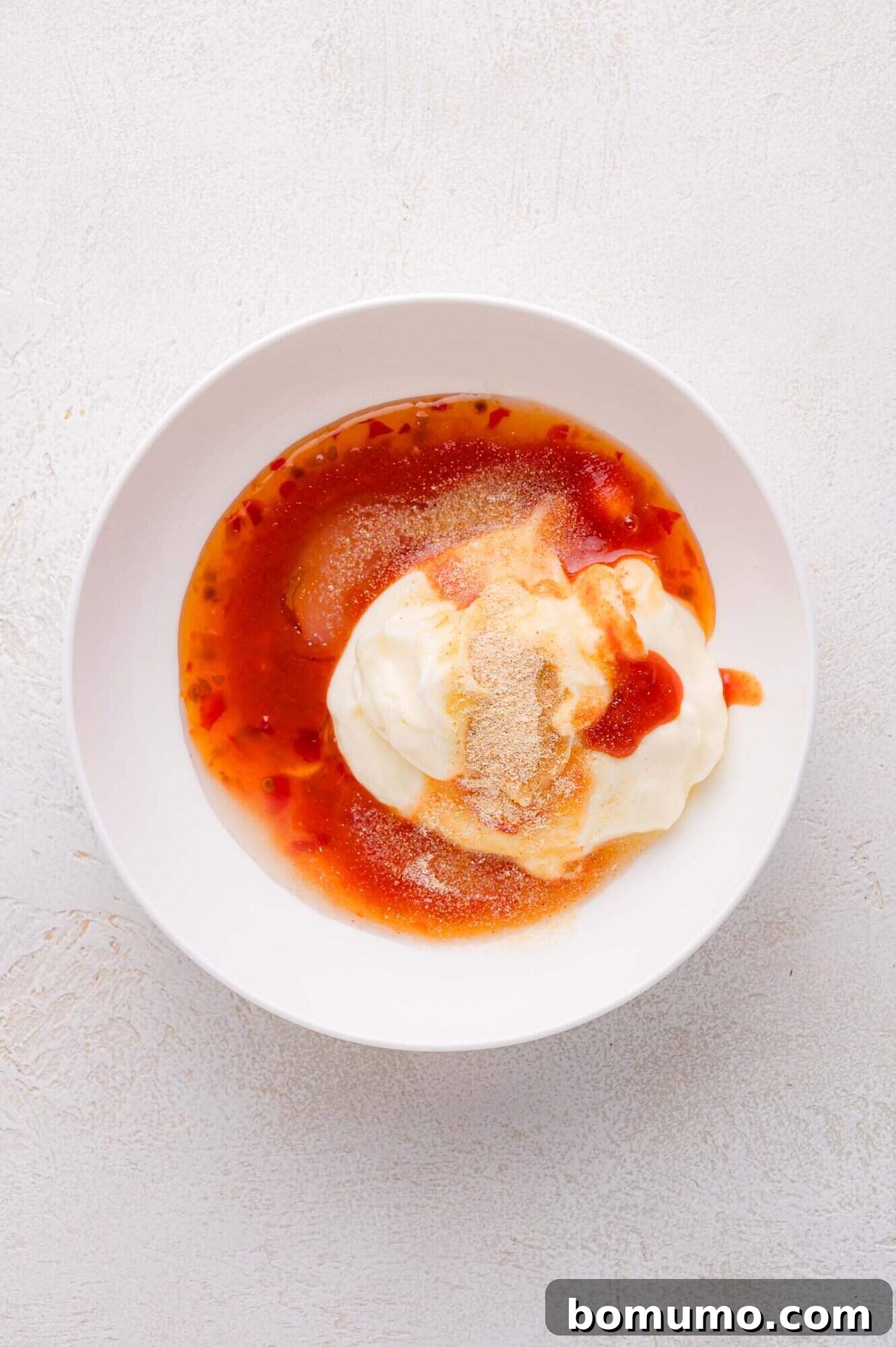 Sweet Chili Blast 5 A crisp white bowl holds a vibrant mixture of creamy mayonnaise, rich sweet chili sauce, and various seasonings, all resting on a subtly textured light surface, poised for whisking into Bang Bang Sauce.
