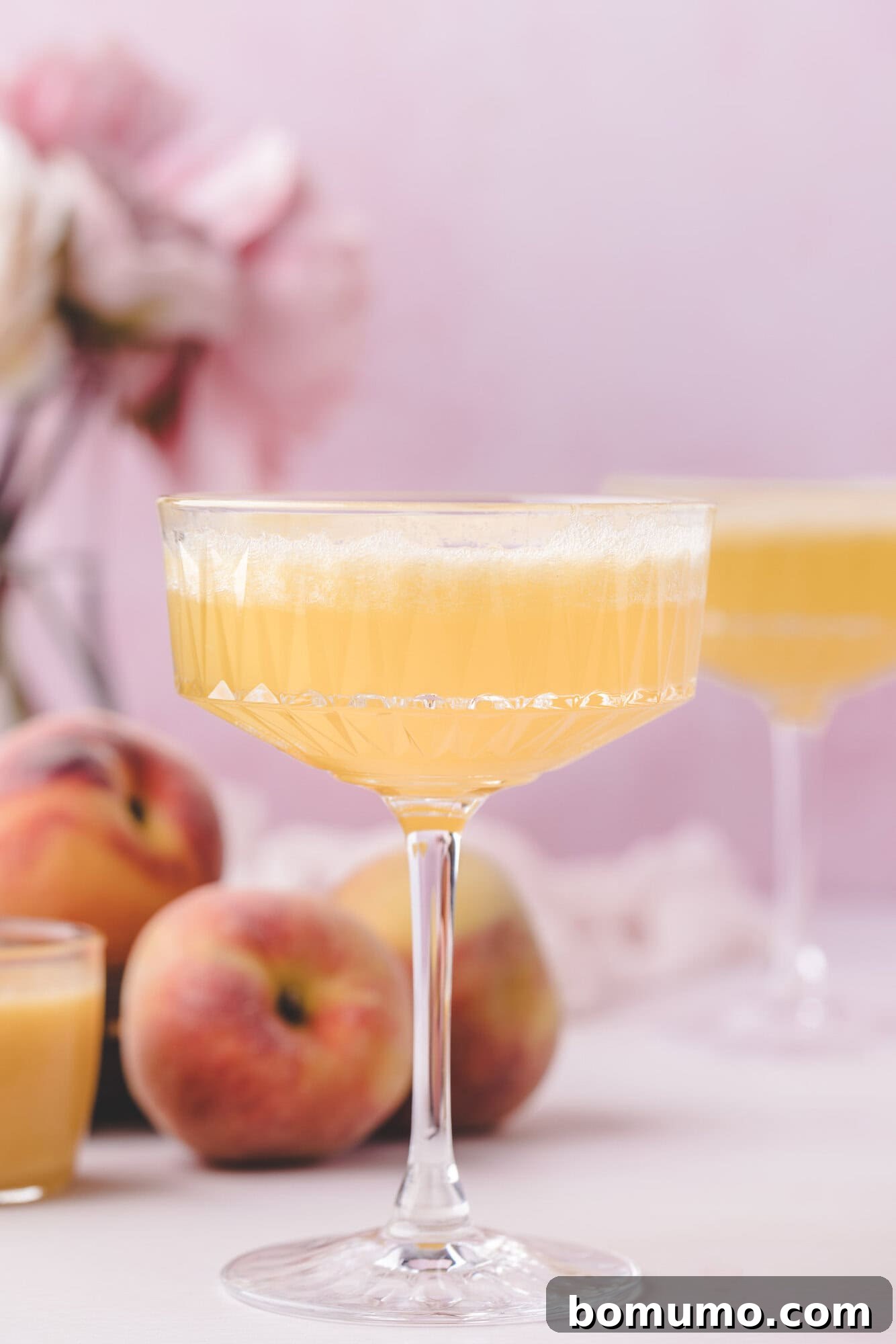 Sparkling Peach Bliss 2 A coupe glass filled with a light orange peach bellini is in focus, with fresh peaches and a blurred pink floral arrangement in the background.