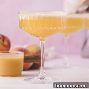 A coupe glass filled with a light orange cocktail, with peaches and another glass in the background on a light pink surface.