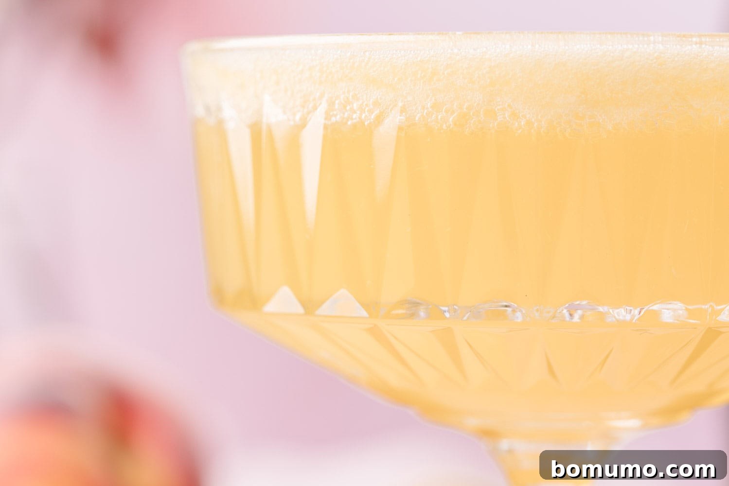 Sparkling Peach Bliss 3 Close-up of a coupe glass filled with a light yellow, frothy beverage against a soft, blurred background.