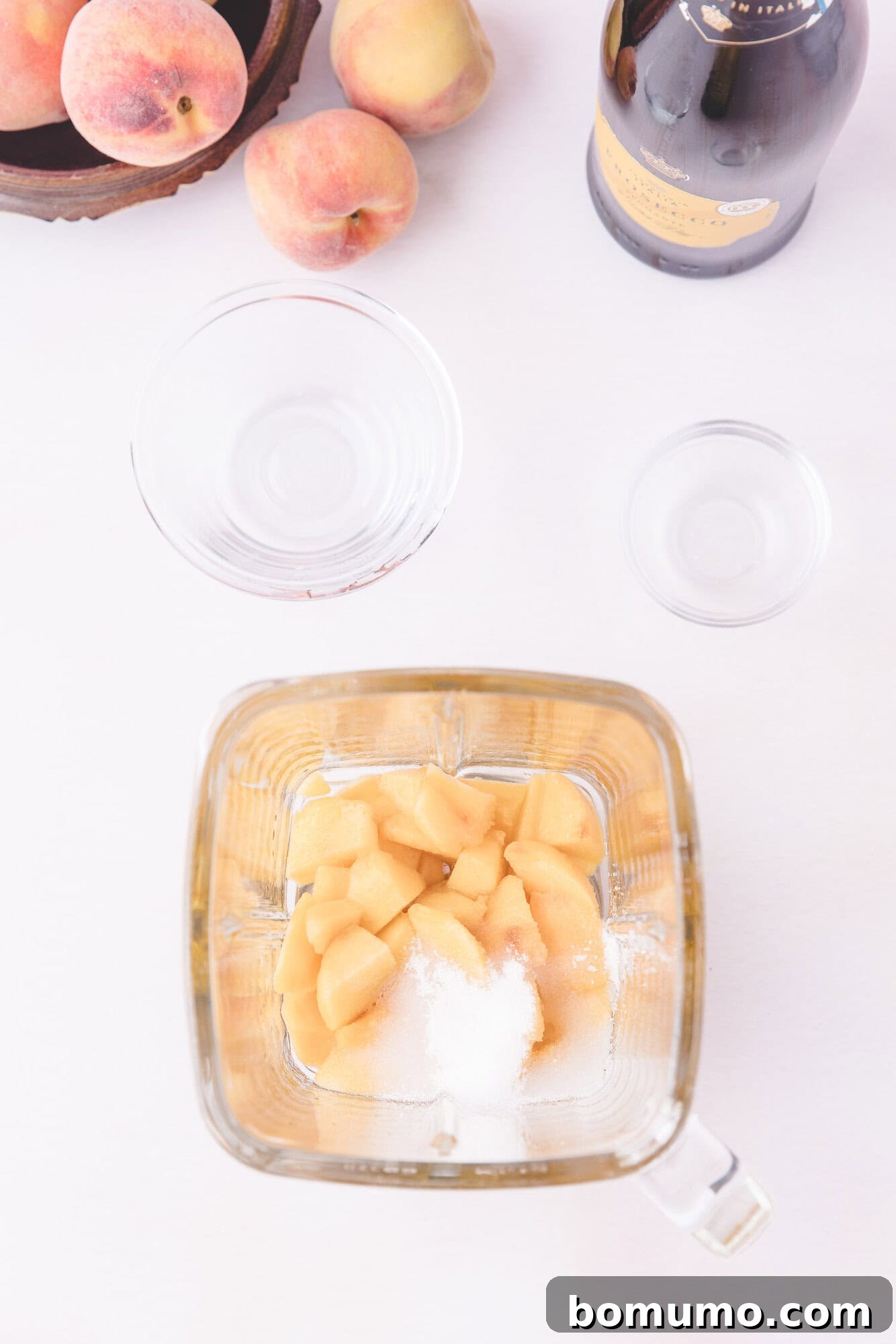 Sparkling Peach Bliss 4 A blender contains chopped peaches and sugar. Nearby are empty glass bowls, a bottle of sparkling wine, and a bowl of whole peaches on a white surface.