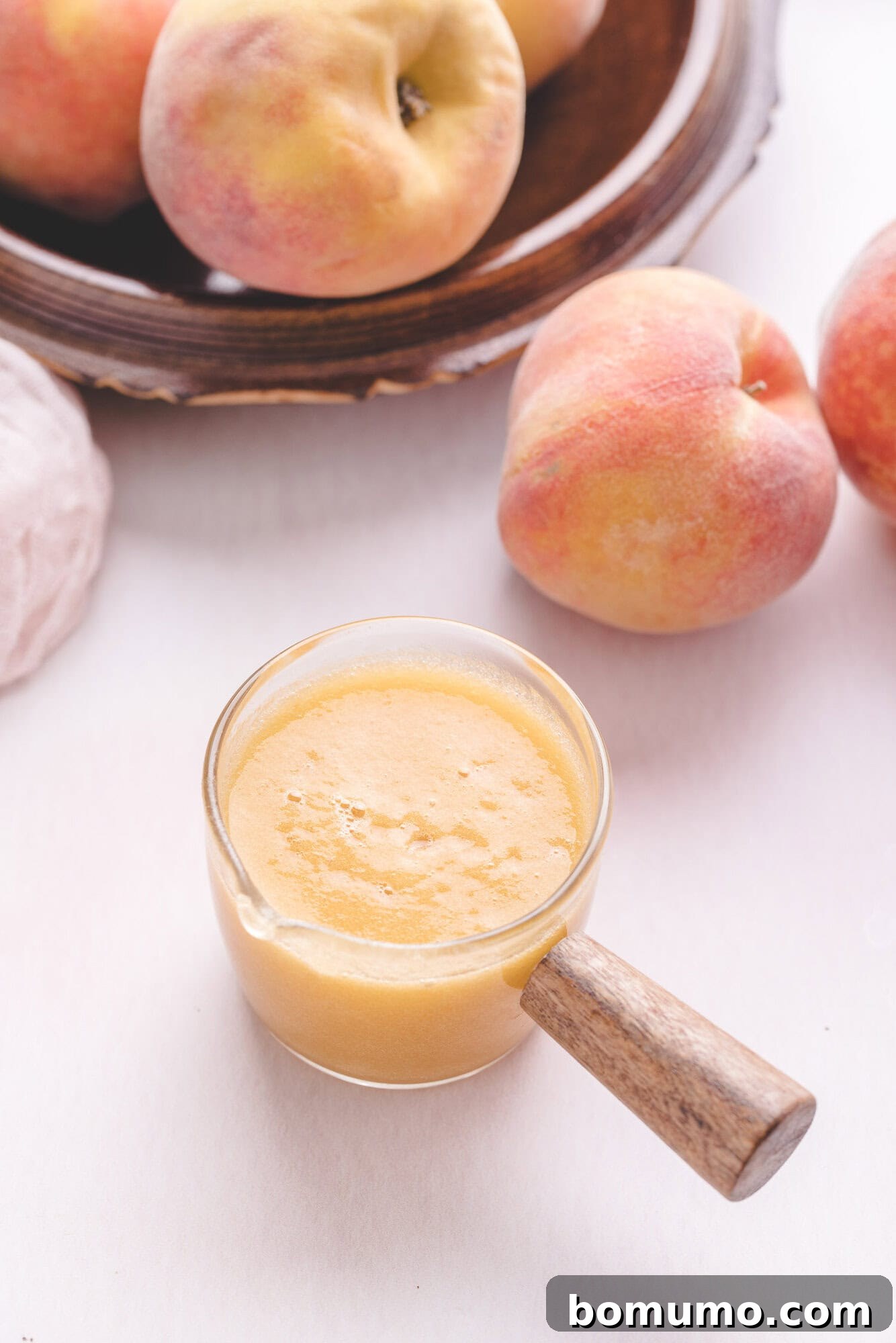 Sparkling Peach Bliss 5 A glass container filled with peach smoothie sits on a white surface, with whole peaches and a wooden plate in the background.