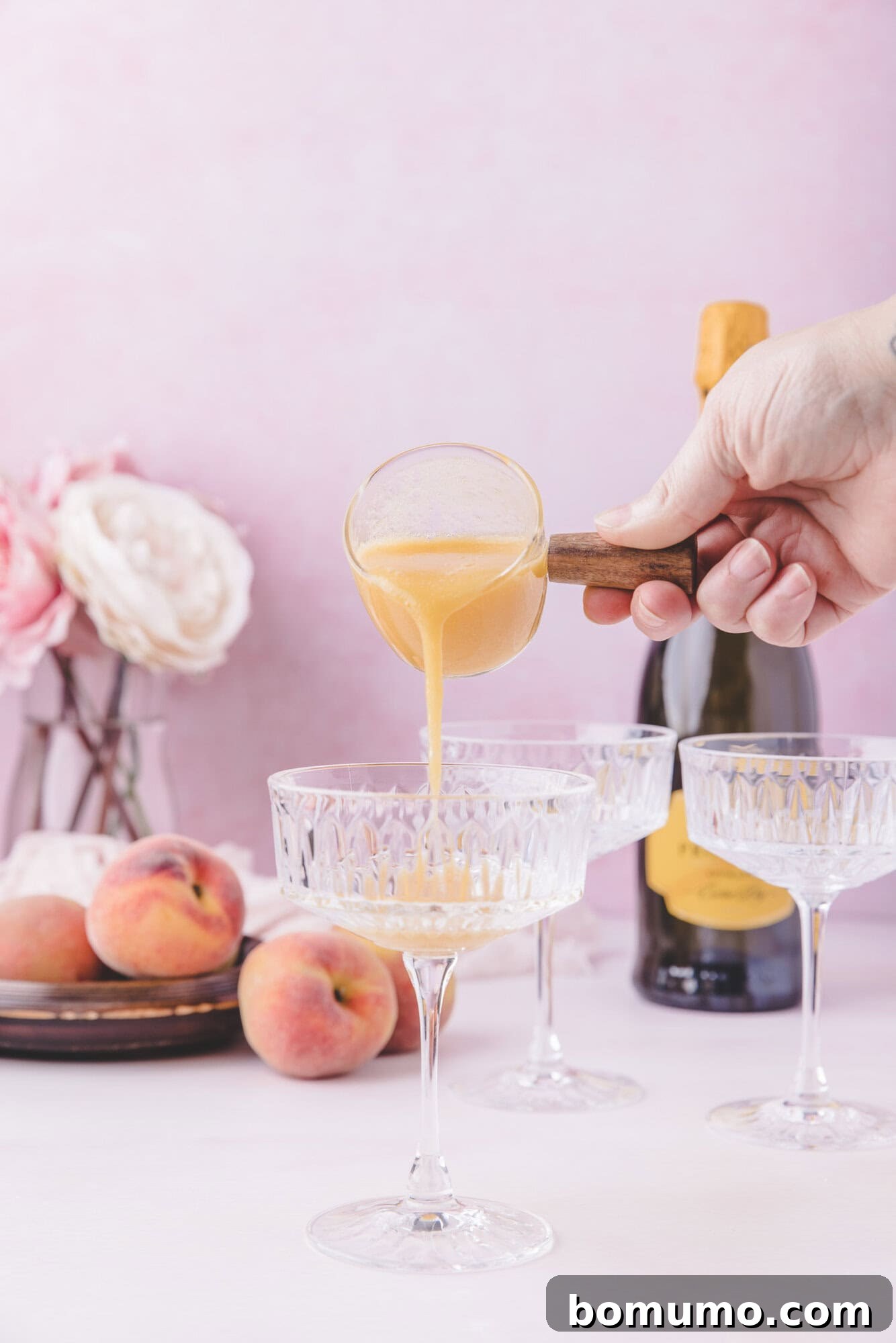 Sparkling Peach Bliss 6 A hand pours peach puree into a coupe glass, surrounded by fresh peaches, a bottle of sparkling wine, and additional empty glasses on a pink background.
