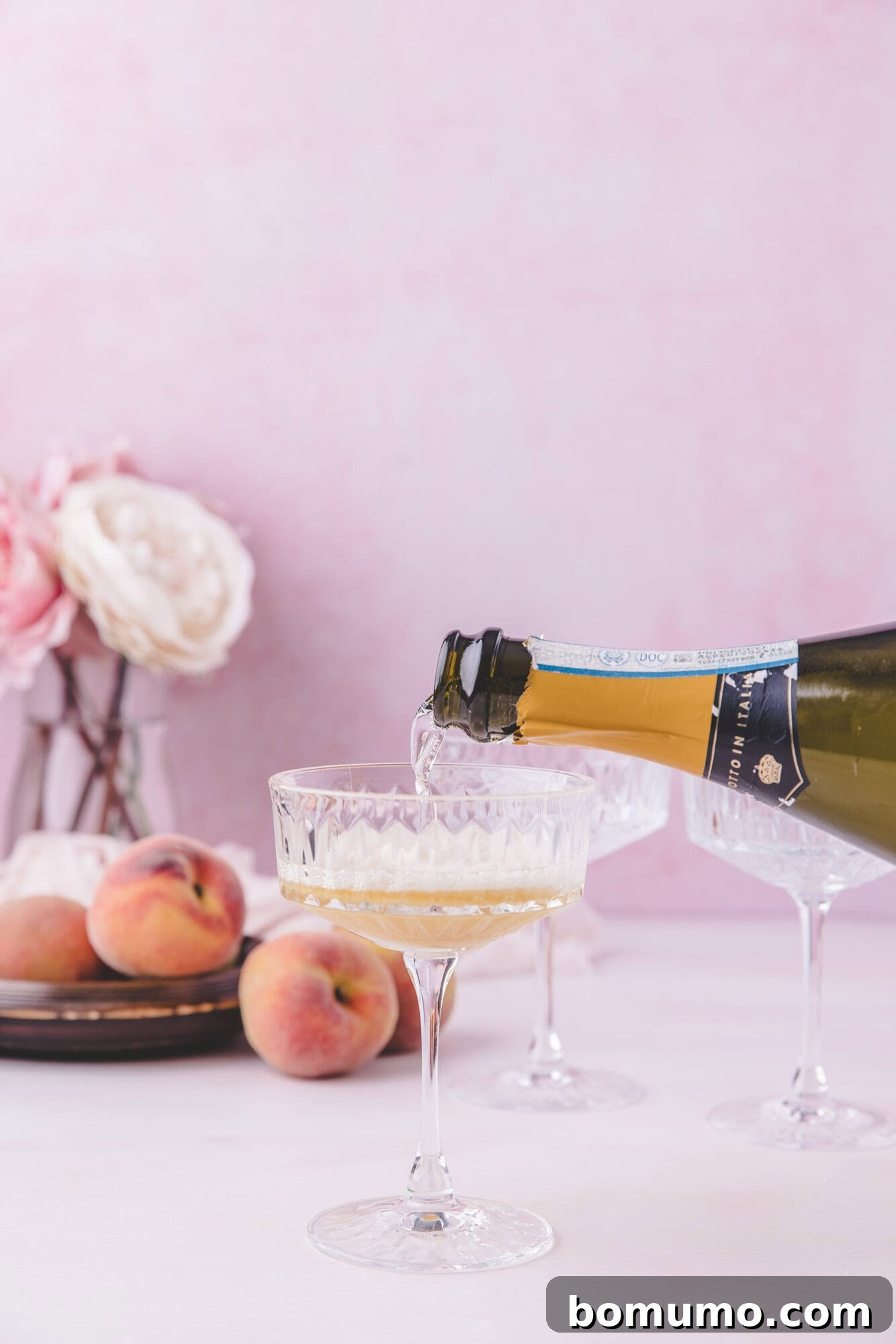 Sparkling Peach Bliss 7 A bottle of sparkling wine is being poured into a coupe glass, with peaches and flowers arranged on a table in the background.