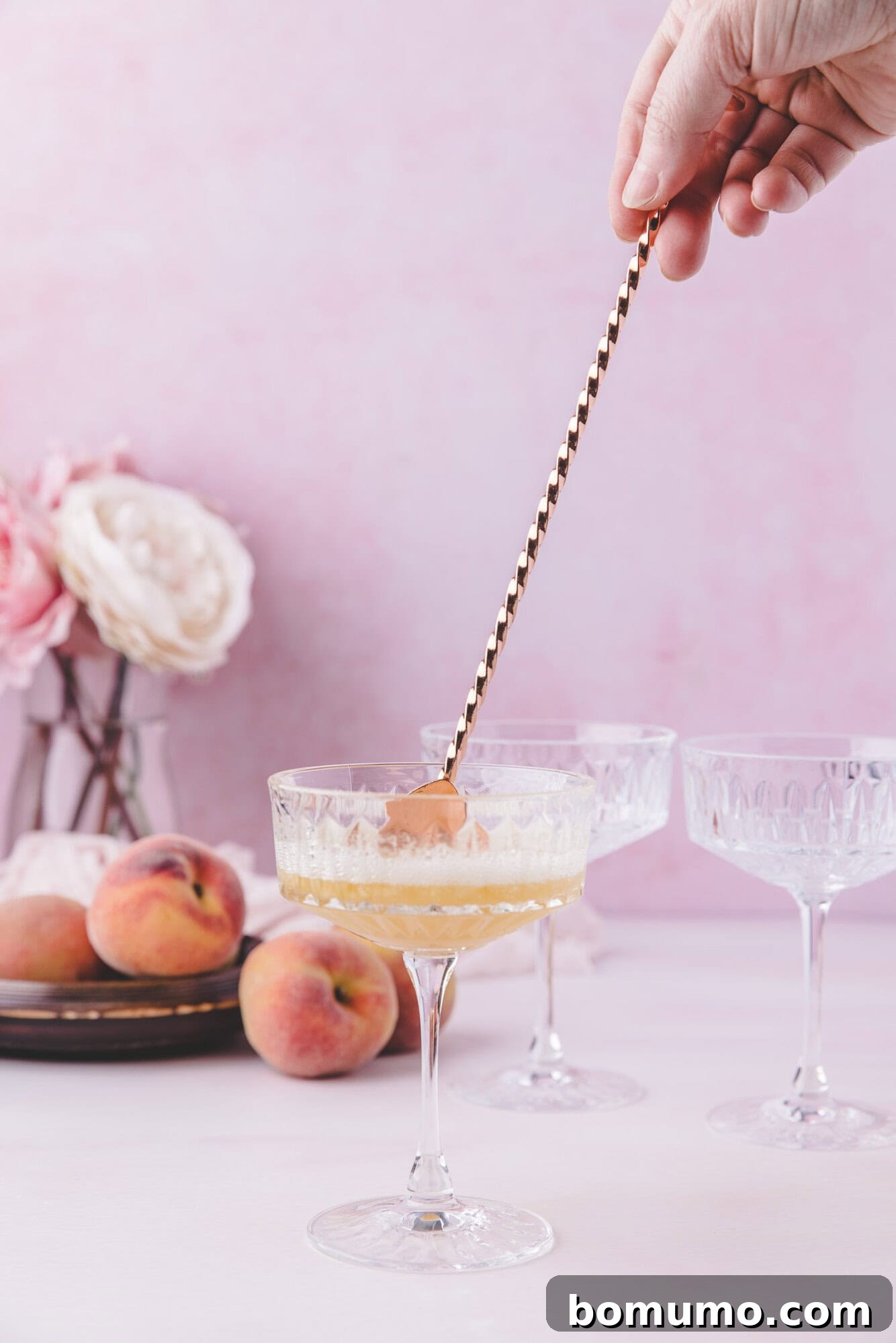 Sparkling Peach Bliss 8 A hand stirs a drink in a coupe glass with a long spoon; peaches and flowers are arranged on a plate in the background.