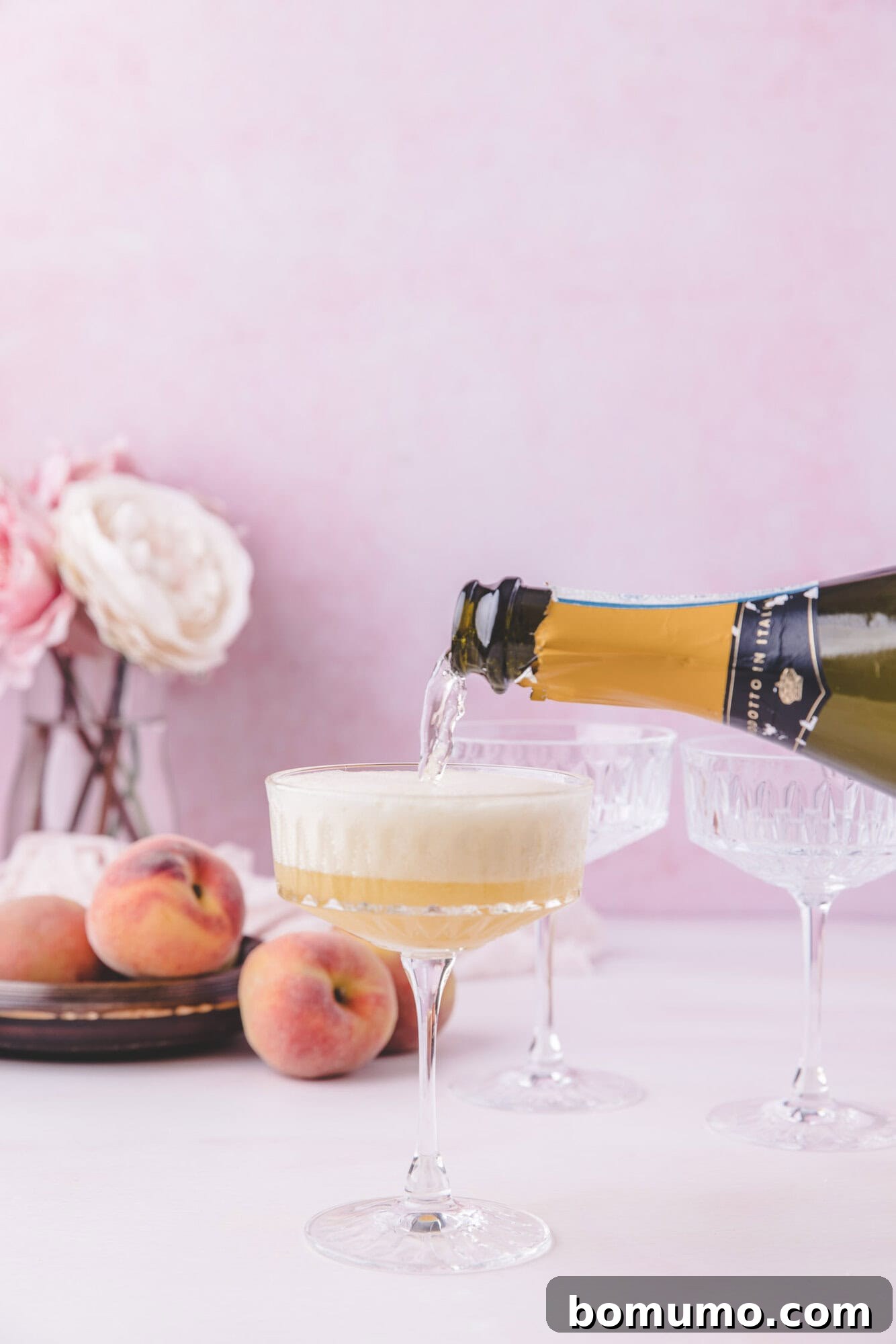 Sparkling Peach Bliss 9 A bottle of sparkling wine is being poured into a coupe glass, with peaches and flowers in the background on a pink surface.
