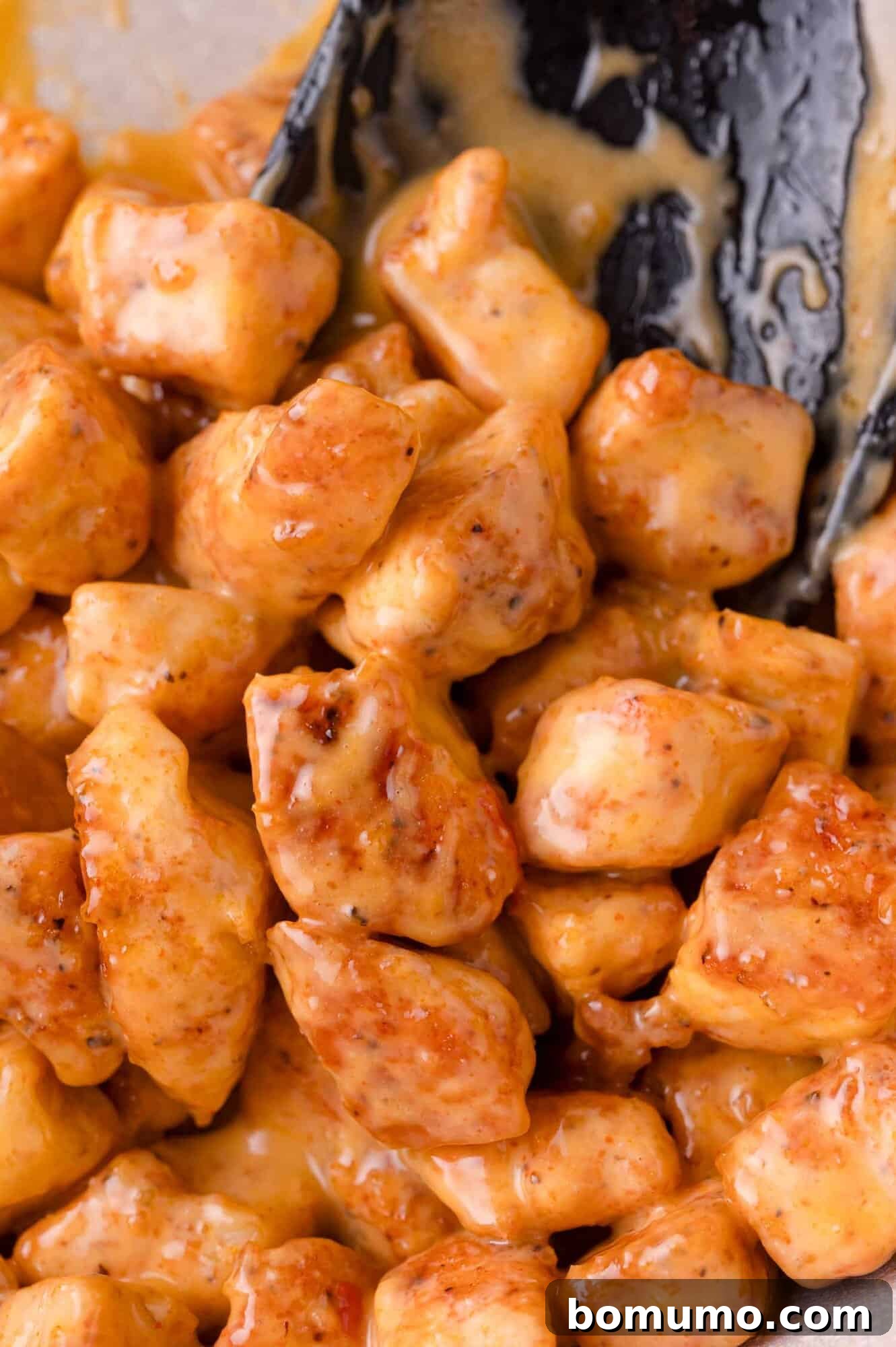 Close-up of golden-brown, bite-sized chicken pieces generously coated in luscious Bang Bang sauce, with a serving utensil partially visible.