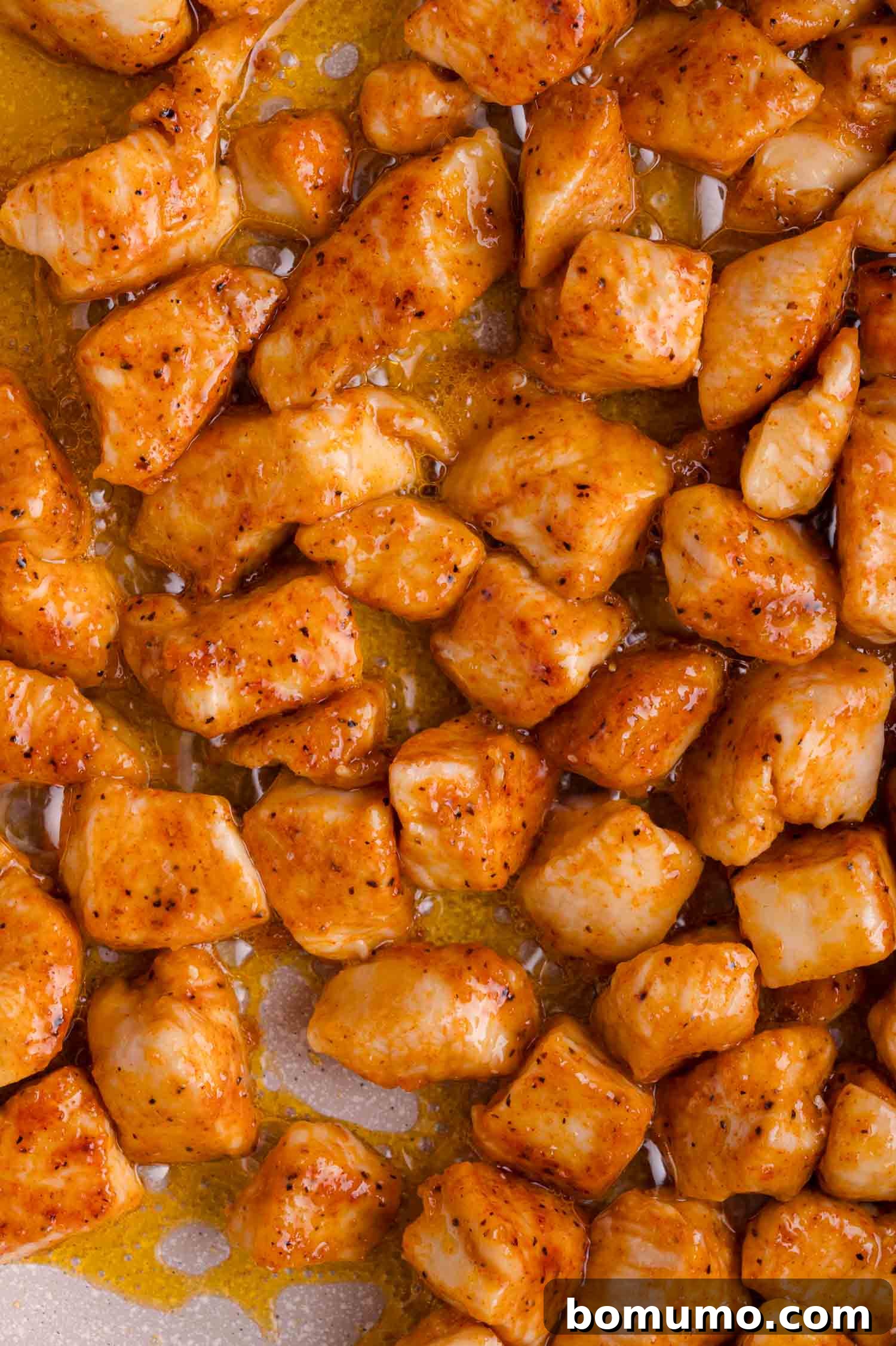 Cooked, seasoned chicken breast pieces beautifully coated in a glossy, orange-brown sauce, ready to be served on a clean white surface.