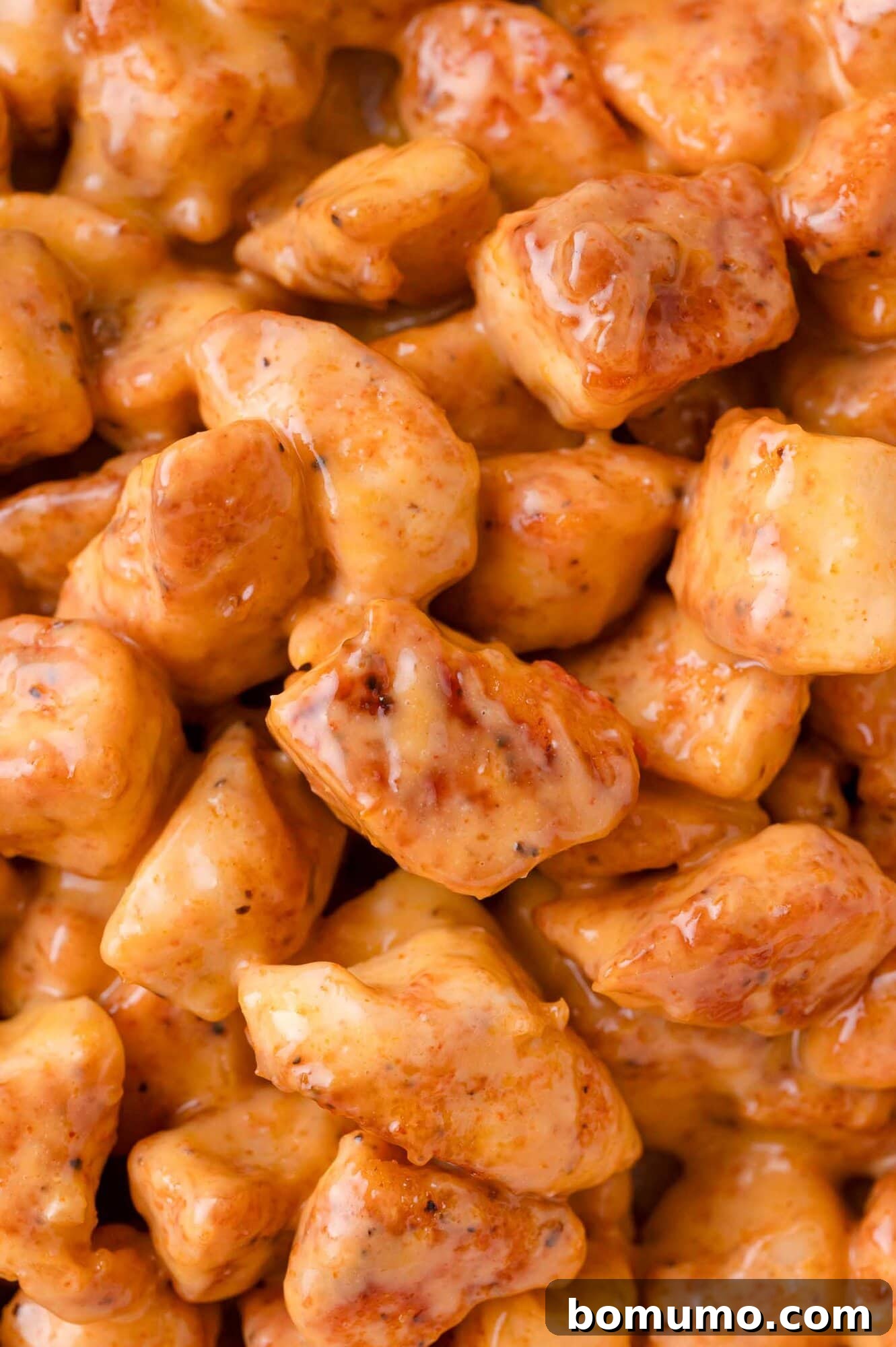 A close-up view of succulent, bite-sized chicken pieces generously coated in a creamy, vibrant orange sauce, highlighting the rich texture.