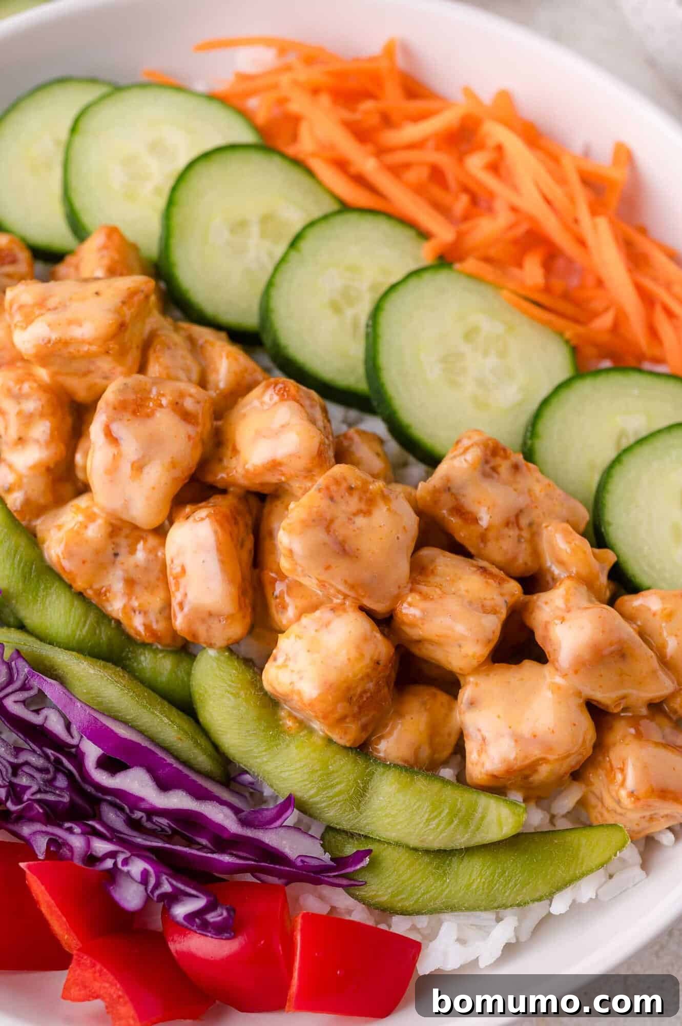 A vibrant bowl featuring fluffy white rice topped with generous portions of Bang Bang Chicken, surrounded by fresh, colorful garnishes including sliced cucumbers, shredded carrots, crisp snap peas, shredded purple cabbage, and diced red bell pepper.