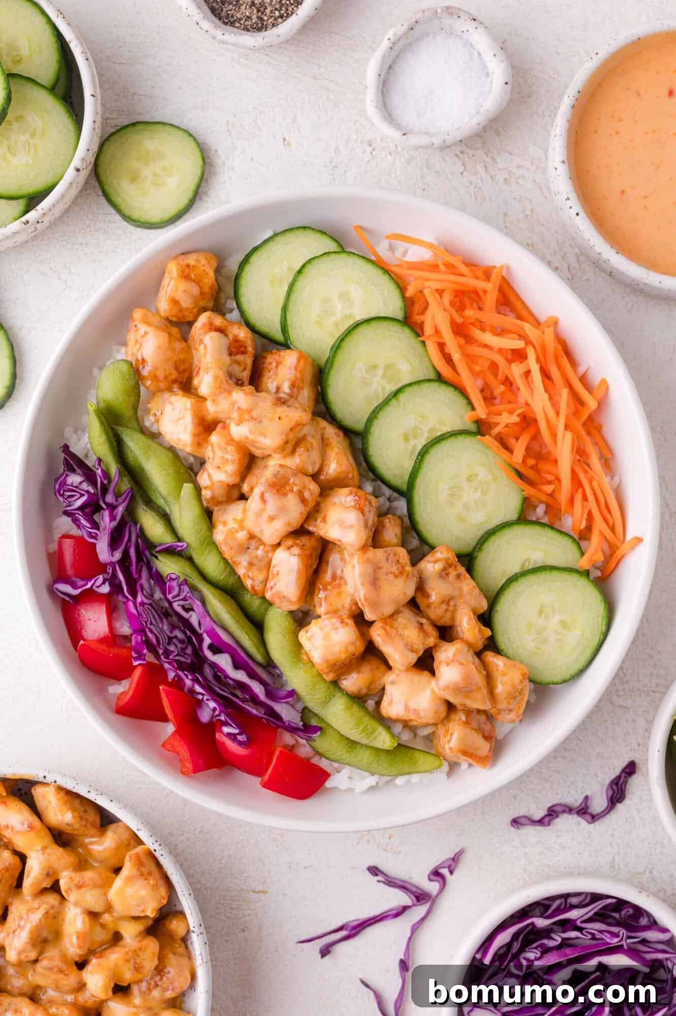 A vibrant and colorful Bang Bang Chicken bowl, served over rice with an assortment of fresh vegetables including sliced cucumber, shredded carrots, red bell pepper, edamame, and purple cabbage. Small bowls of sauce and additional vegetables surround the main dish.