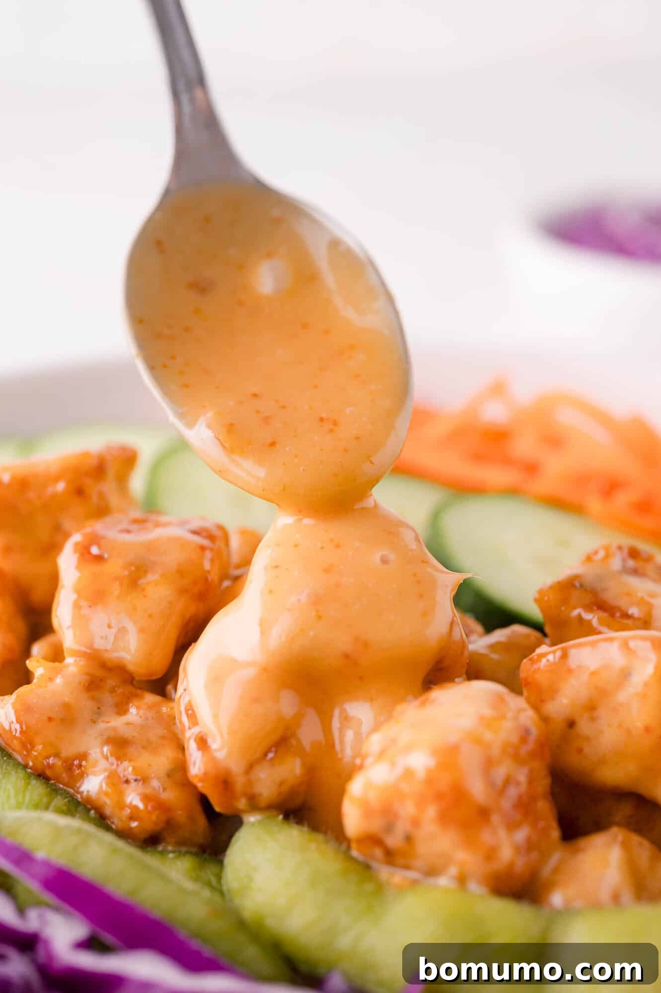 A spoon drizzles creamy, vibrant Bang Bang sauce over a bed of golden-brown chicken pieces, accompanied by fresh, colorful vegetables like sliced cucumber, shredded carrots, and crisp cabbage.