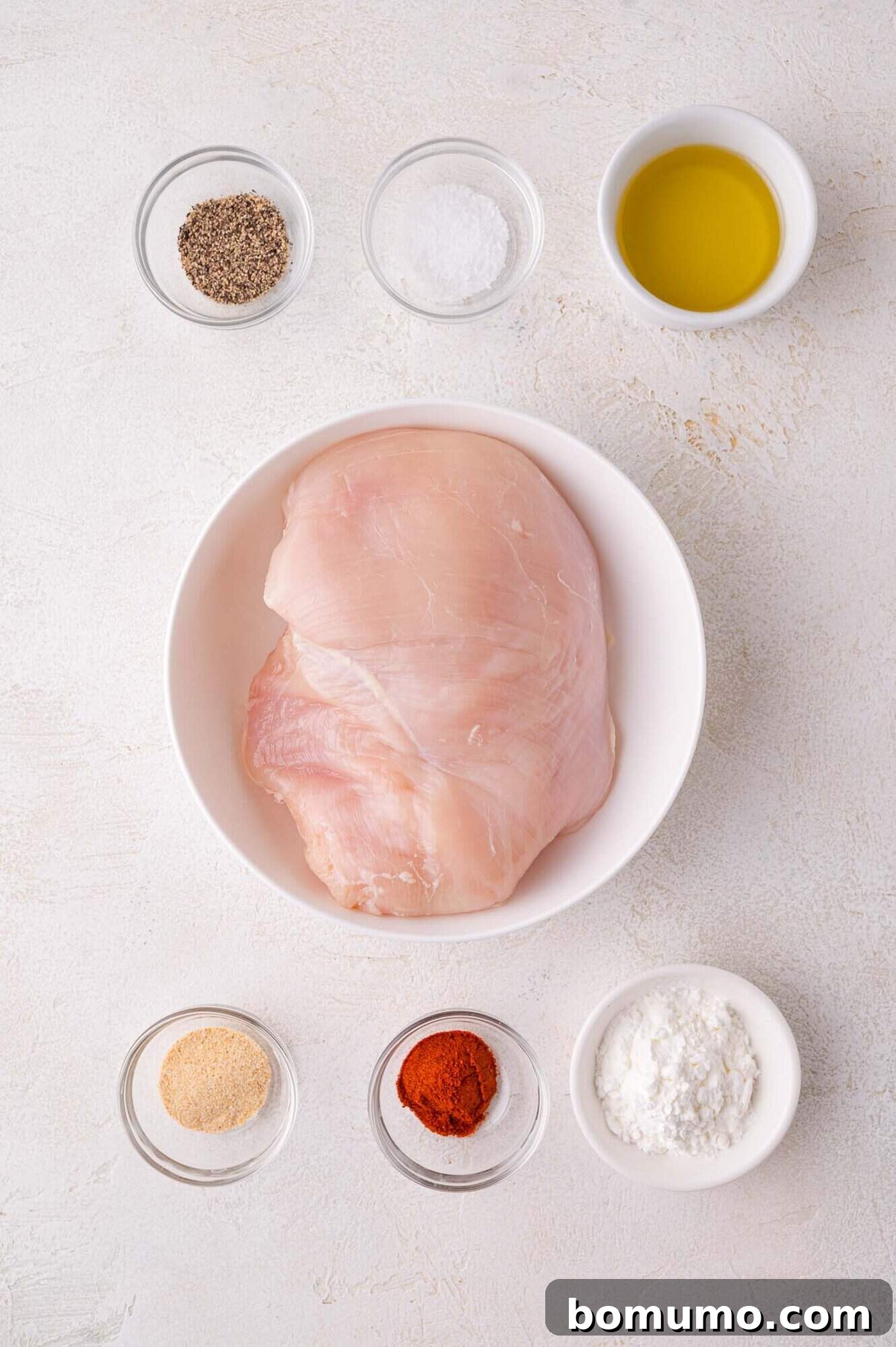 Raw boneless, skinless chicken breast in a white bowl, surrounded by smaller bowls containing the dry seasonings: black pepper, salt, olive oil, garlic powder, paprika, and cornstarch, all on a light surface, ready for preparation.