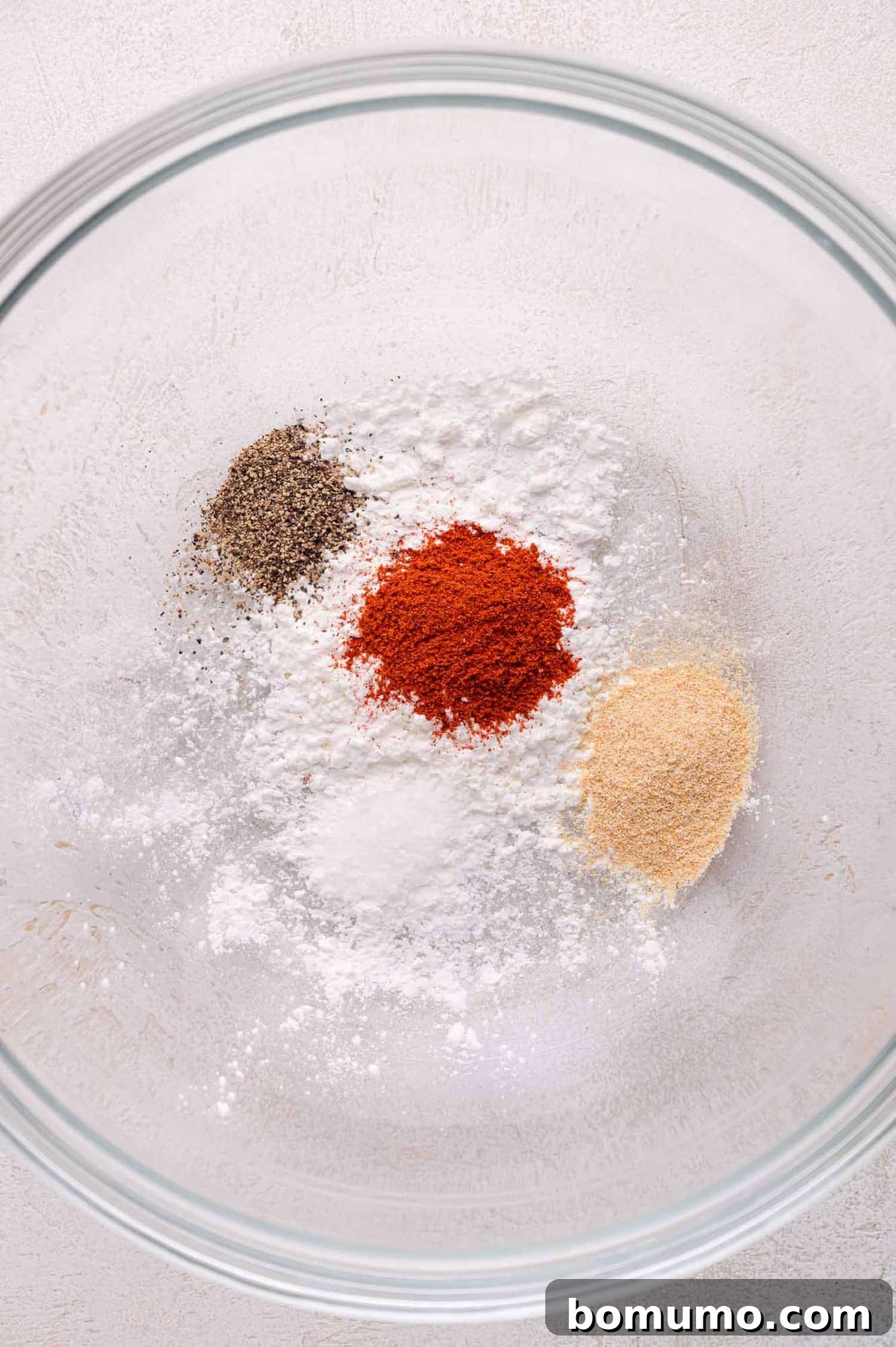 A glass bowl holds separate piles of black pepper, paprika, garlic powder, and cornstarch on a textured light surface, awaiting mixing.