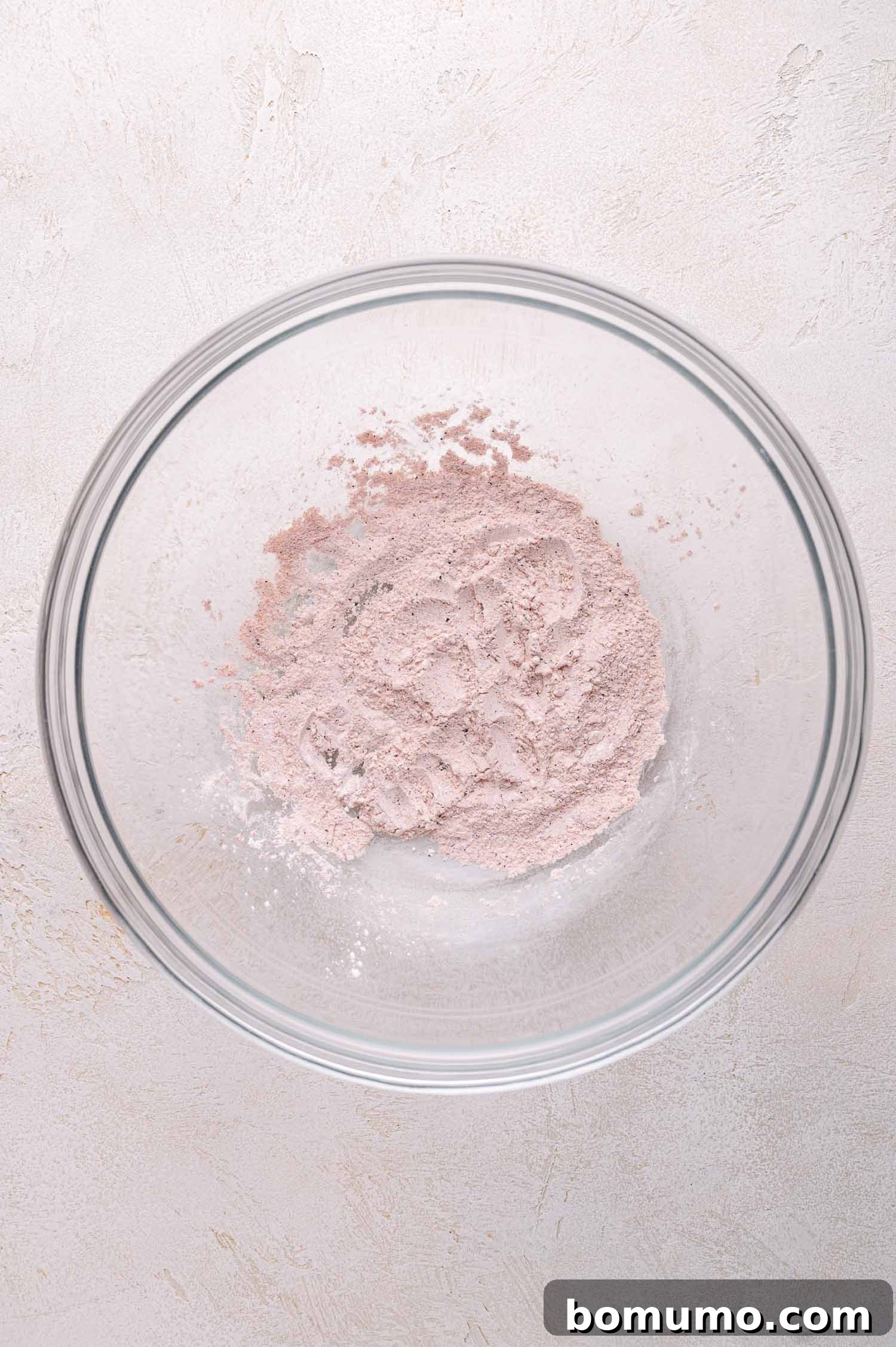 A small amount of light pink seasoned cornstarch mixture settled at the bottom of a clear glass mixing bowl on a light-colored surface.