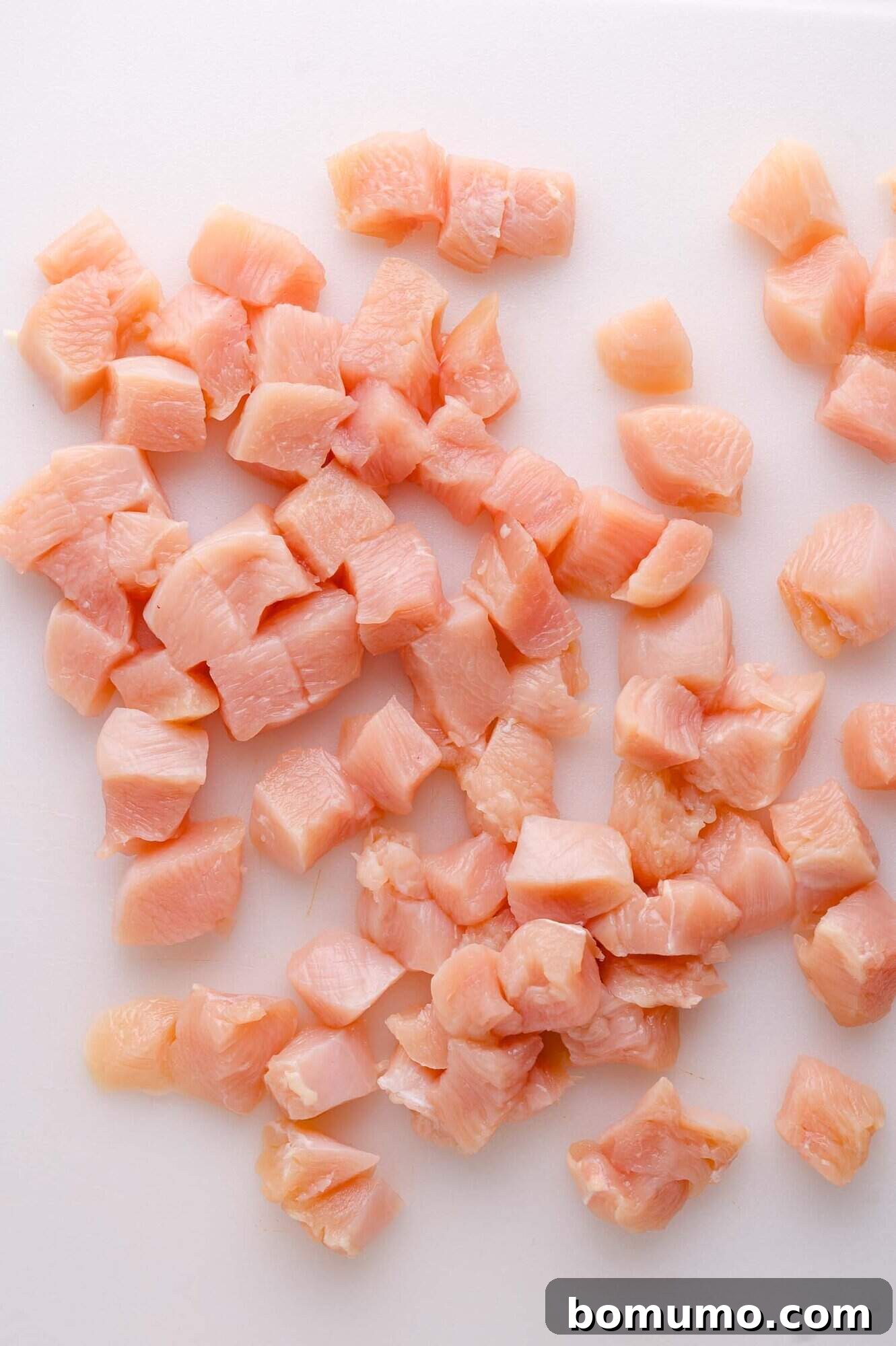 Neatly diced raw chicken pieces are spread out on a clean white cutting board, showcasing uniform size for even cooking.