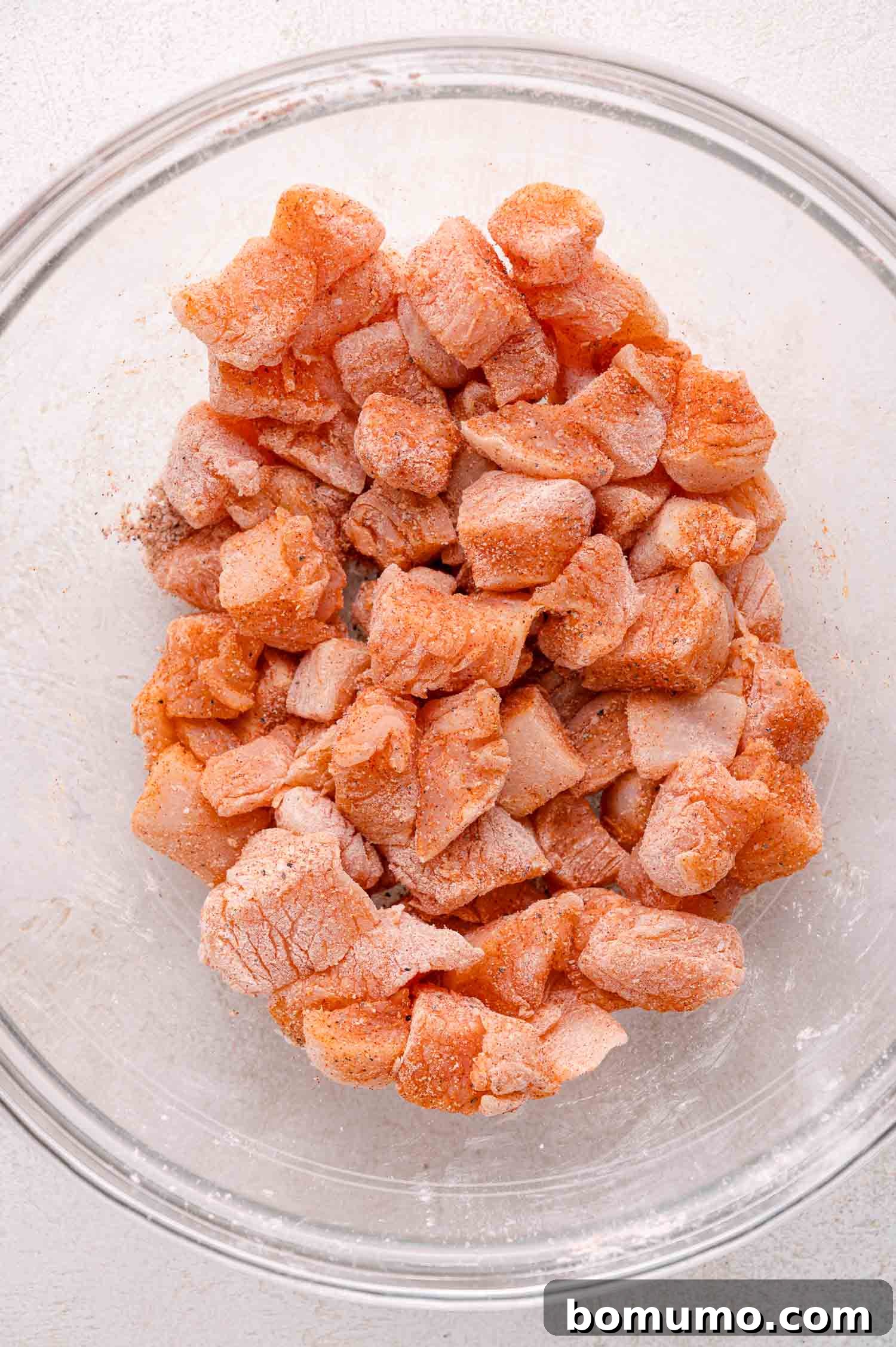Raw, cubed chicken pieces are thoroughly coated with a light blend of spices and cornstarch in a clear glass mixing bowl.