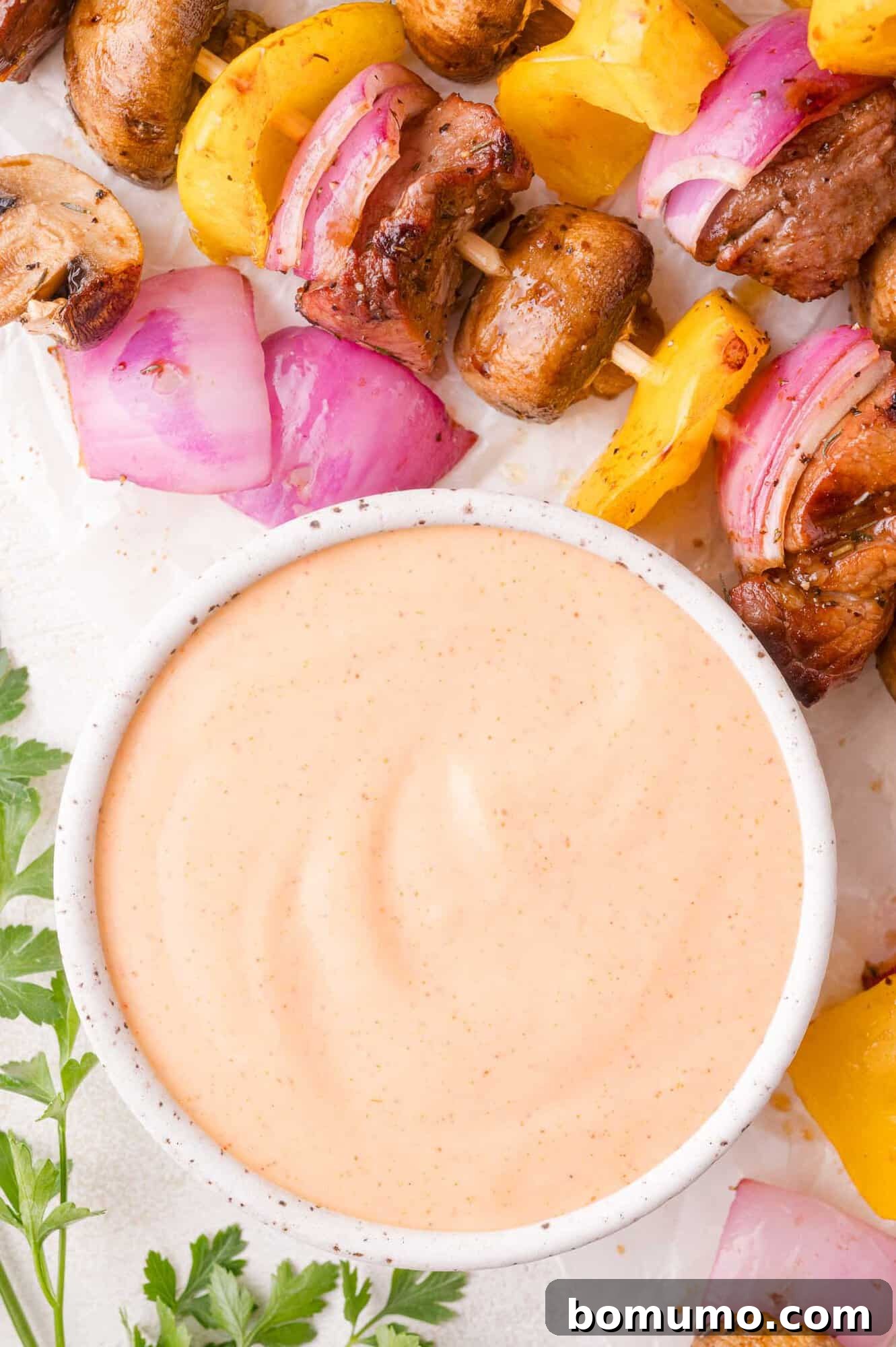A bowl of creamy dipping yum yum sauce sits next to grilled vegetable and meat skewers with mushrooms, red onions, and yellow bell peppers. Parsley is visible in the corner.