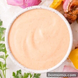 A white bowl filled with creamy, light orange yum yum sauce, surrounded by chopped parsley, onions, and yellow bell pepper pieces.