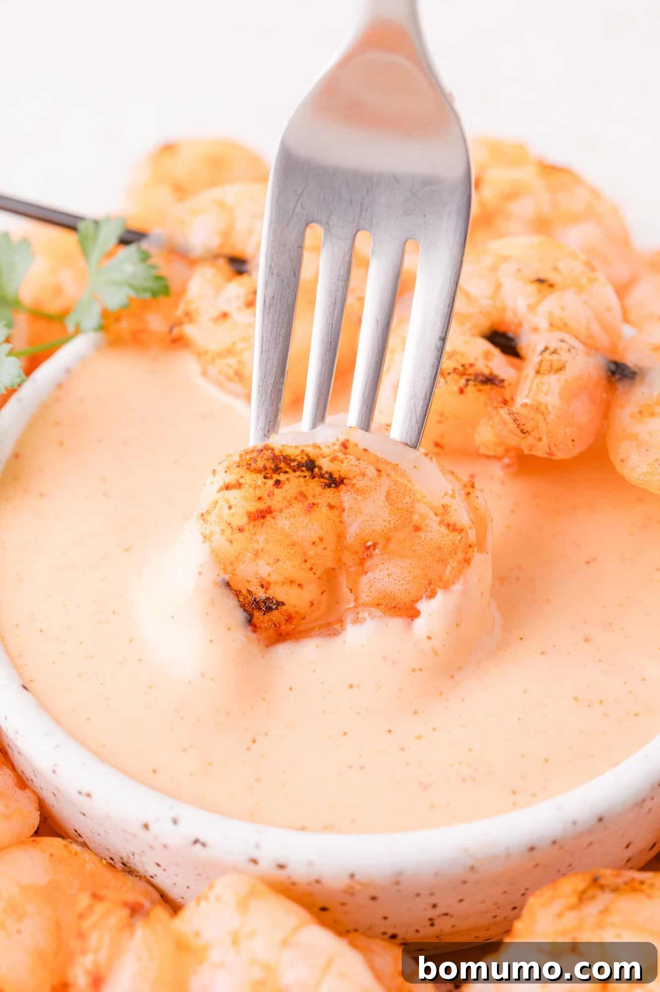 A grilled shrimp held by a fork is being dipped into a bowl of creamy orange sauce, with more shrimp and a sprig of parsley in the background.