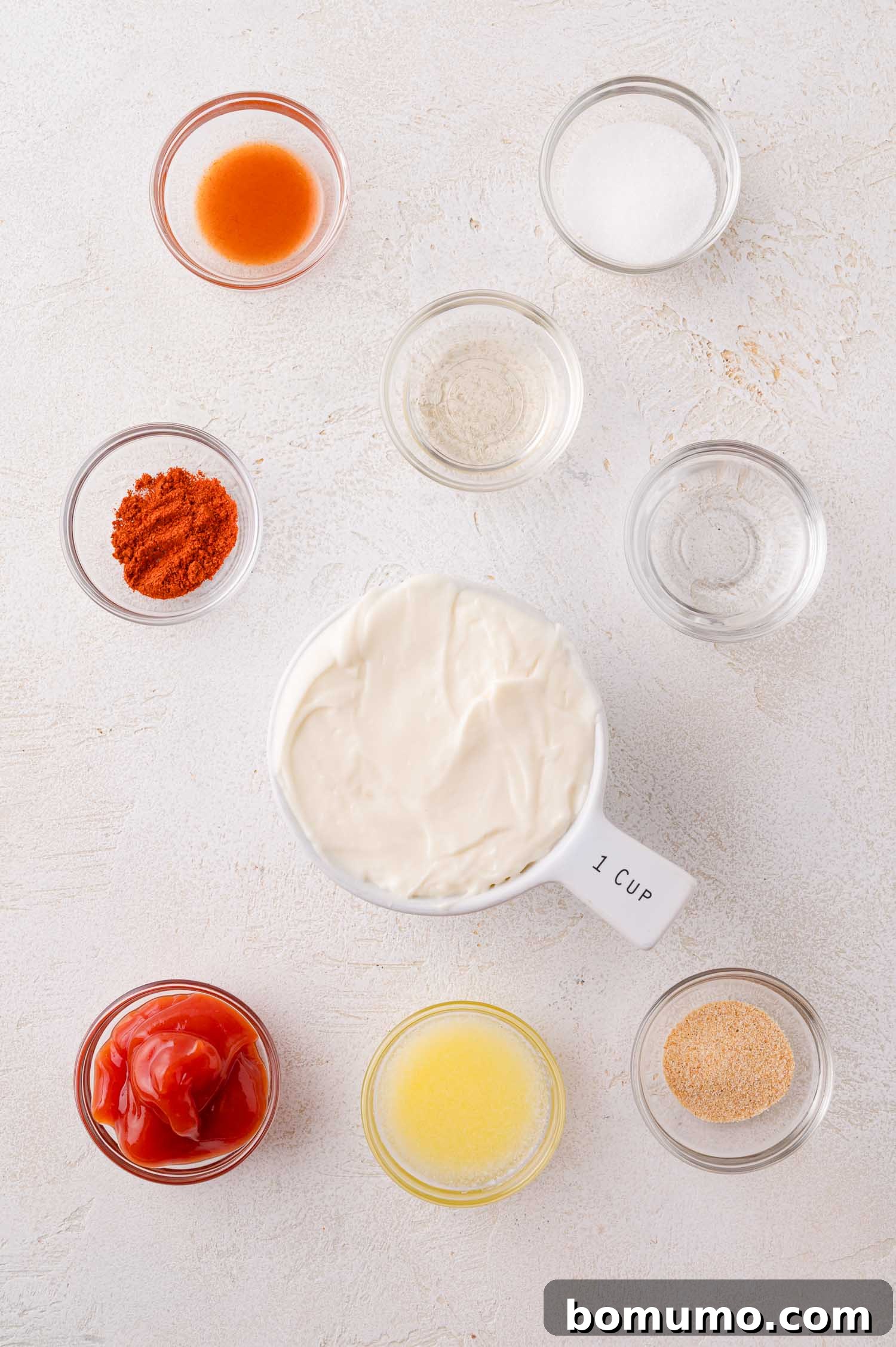 Nine small bowls containing various ingredients, including mayonnaise, ketchup, hot sauce, spices, and liquids, arranged on a white surface.