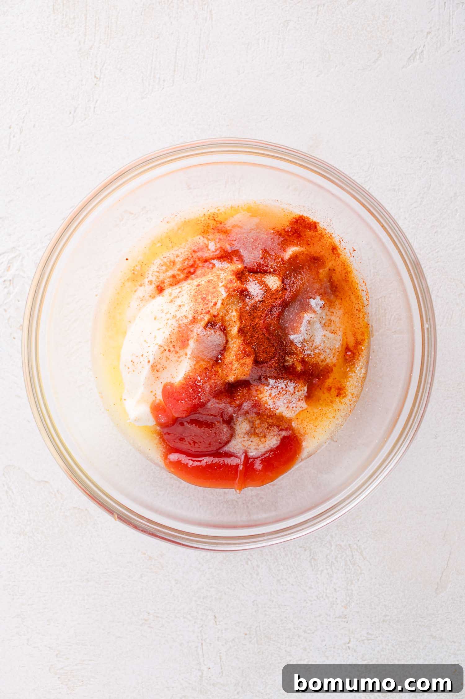 A glass bowl containing mayonnaise, ketchup, and various spices, sitting on a white surface.