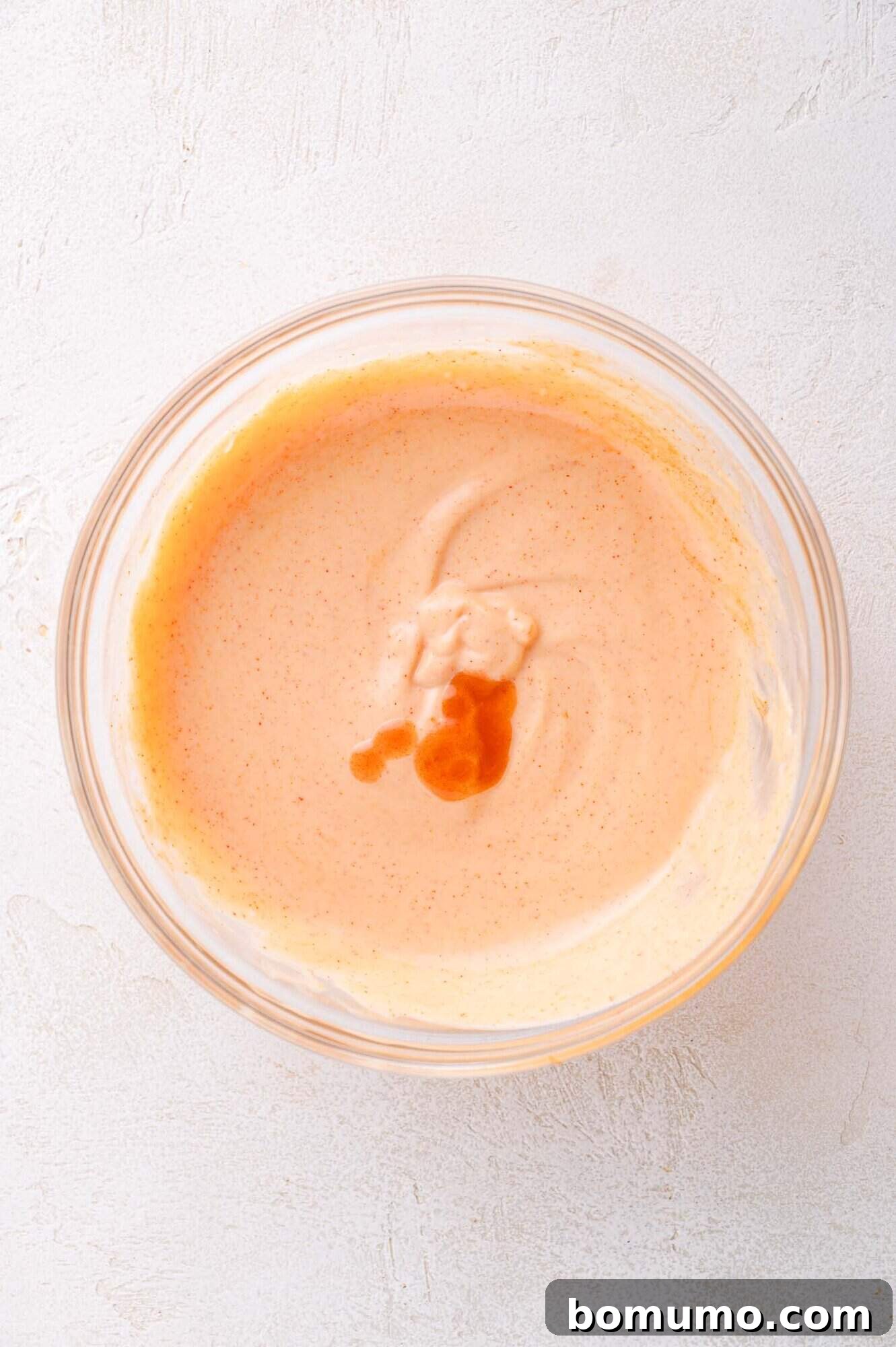 A glass bowl containing a creamy, light orange sauce with a few drops of darker liquid on top, set on a light textured surface.