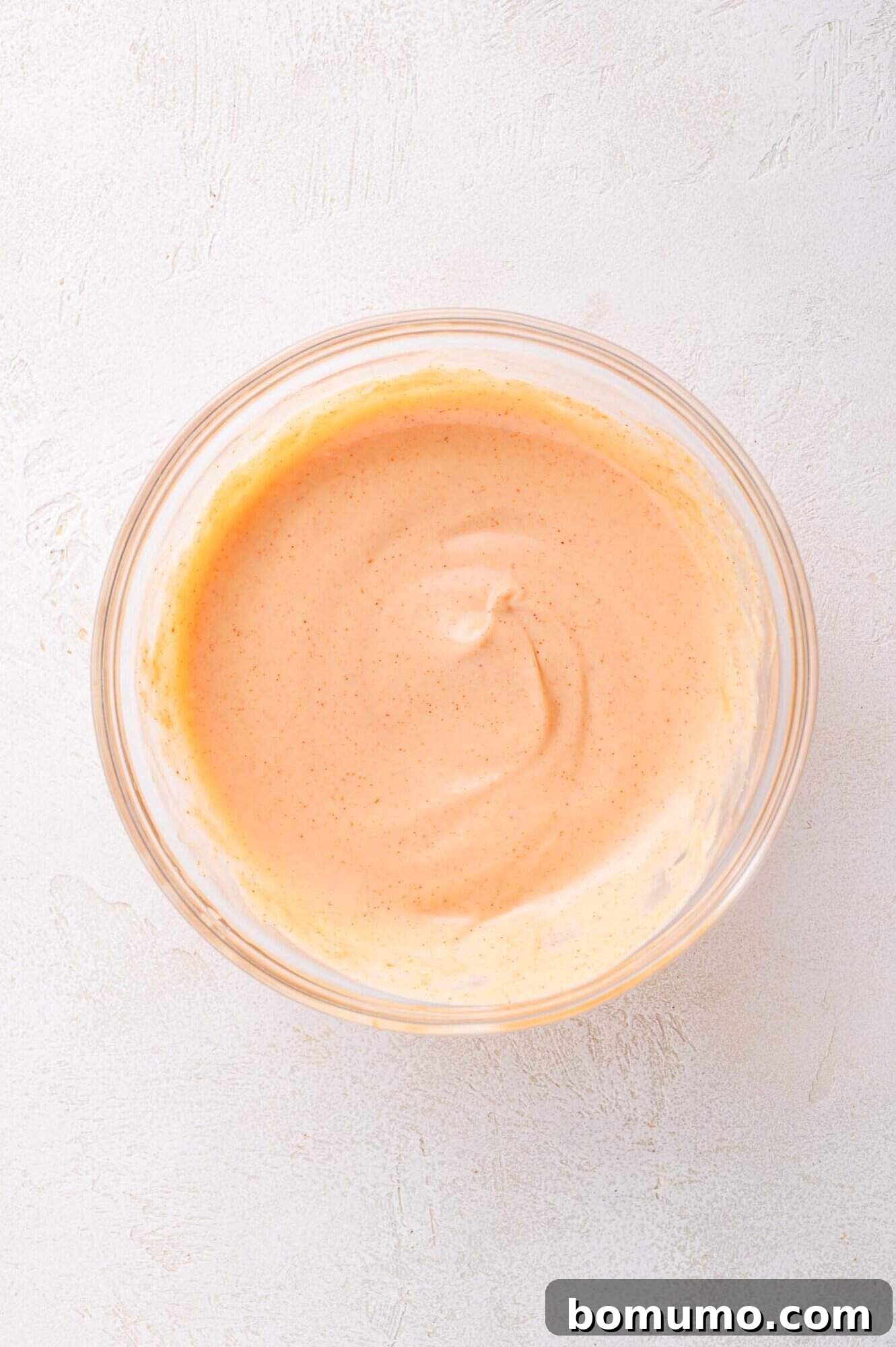 A glass bowl filled with creamy, orange-colored yum yum sauce sits on a light textured surface.