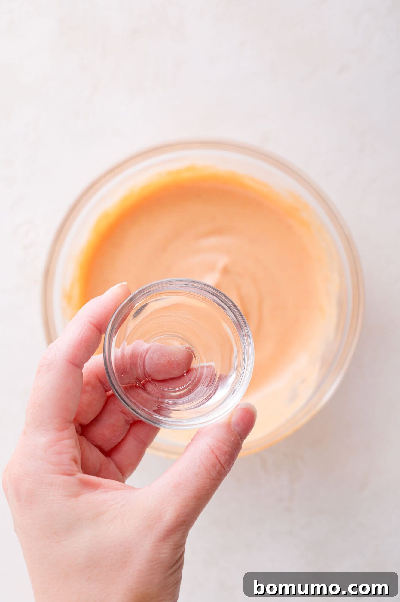 A hand holding a small clear glass bowl above a larger glass bowl containing an orange mixture on a light surface.