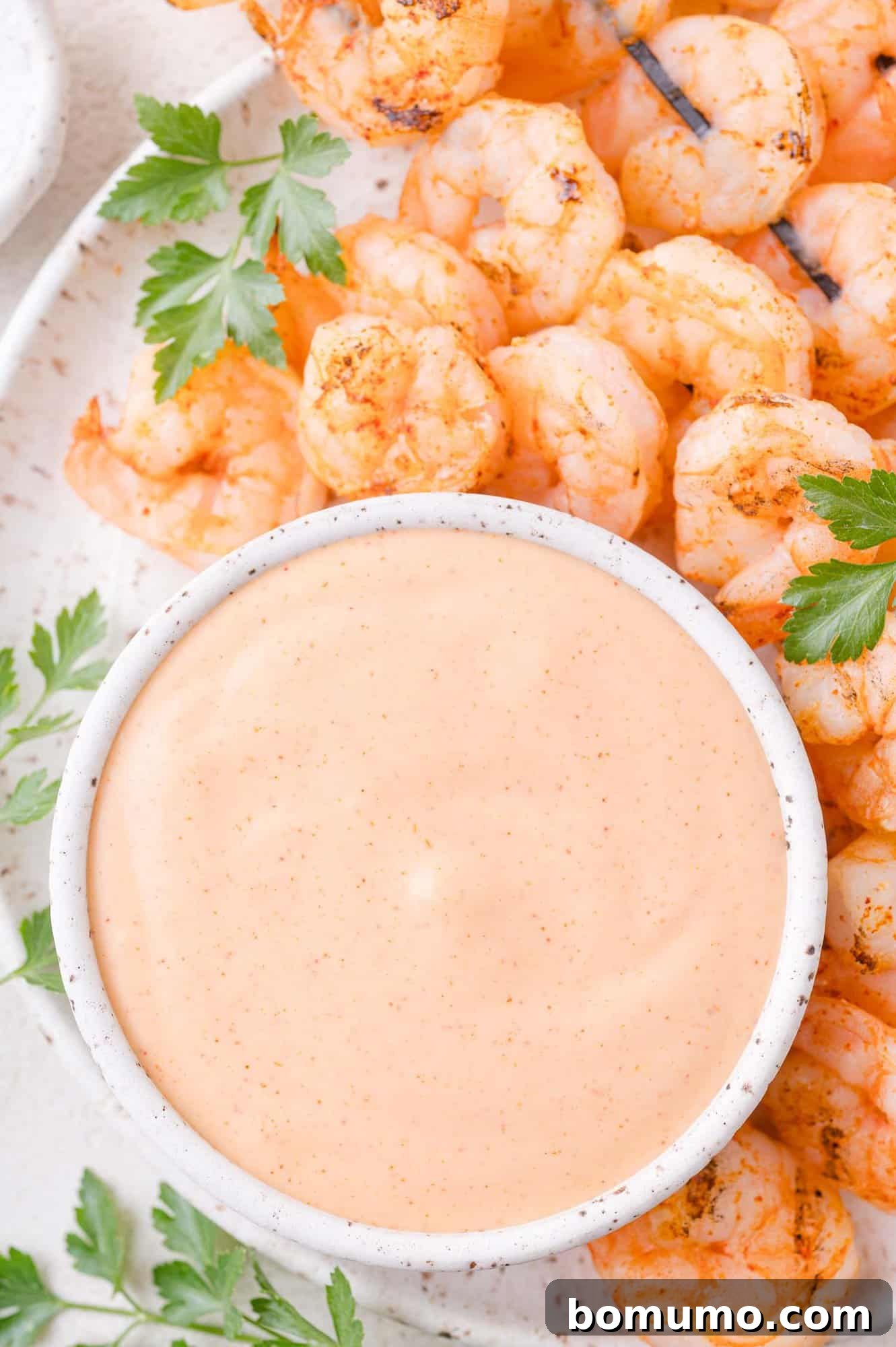 A bowl of creamy orange dipping sauce is surrounded by grilled shrimp and fresh parsley on a white plate.