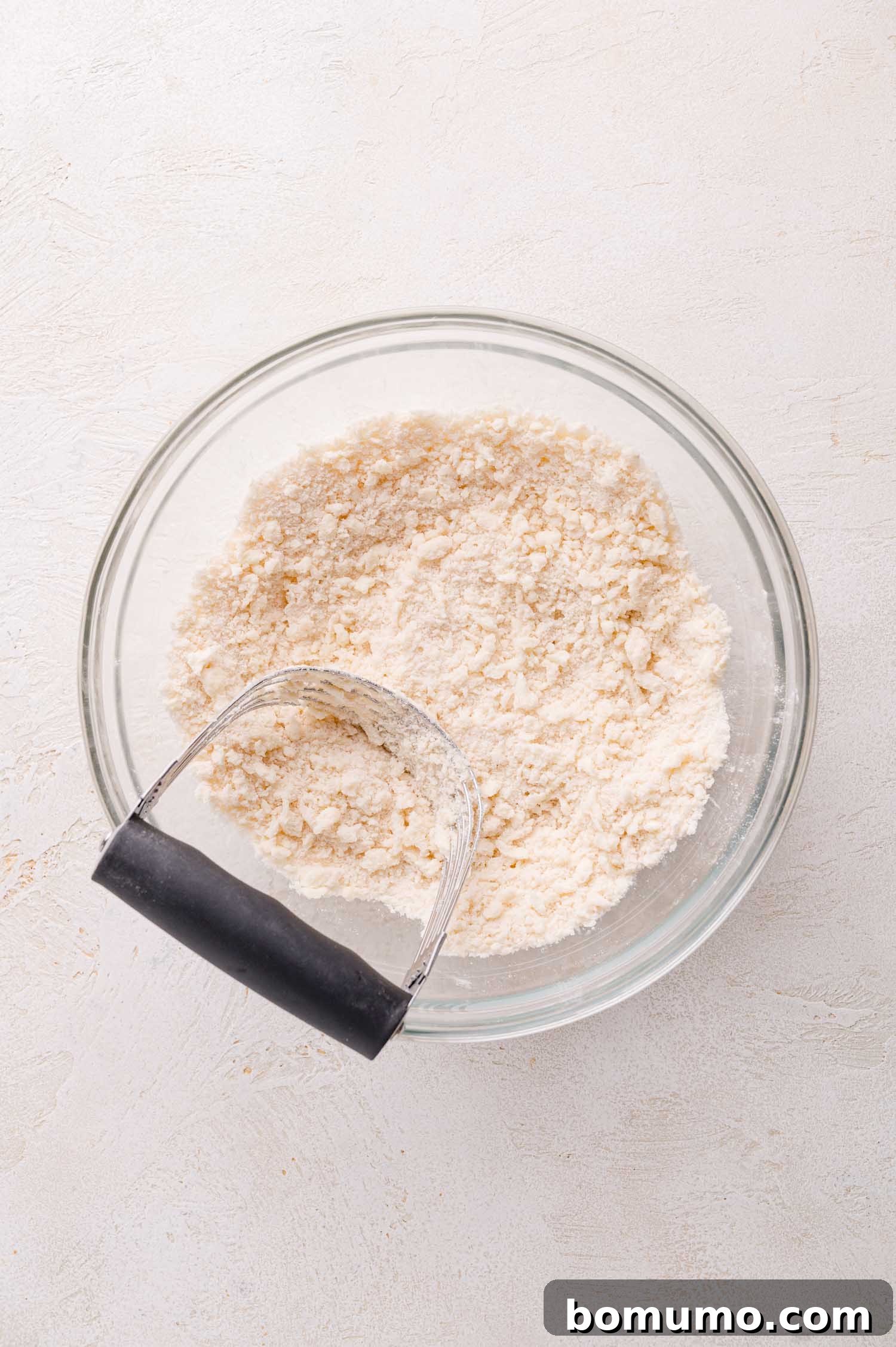 A close-up of a glass bowl holding a coarse, crumbly flour mixture, with a pastry cutter resting inside, illustrating the ideal texture before adding liquid ingredients.