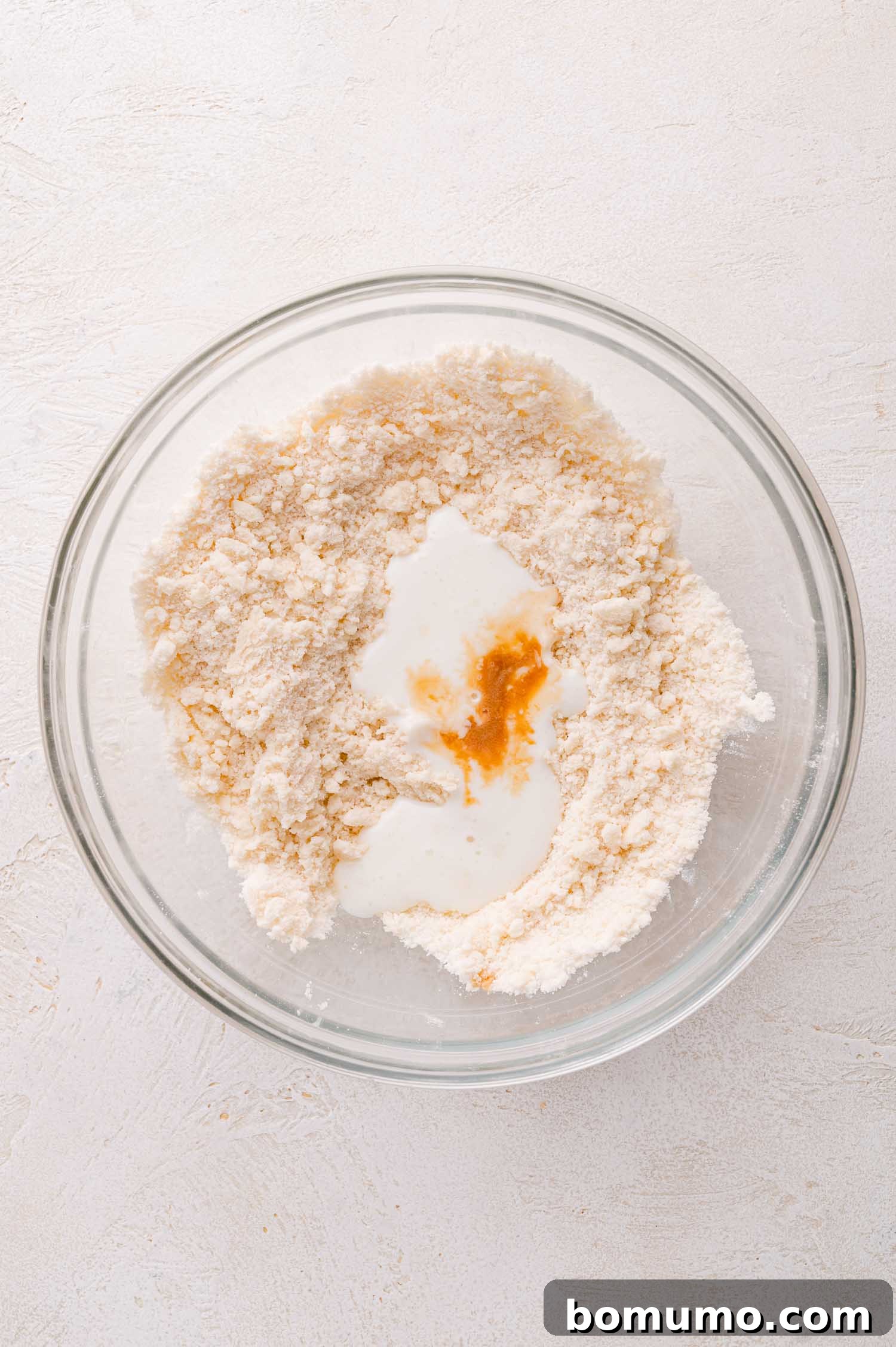 A clear glass bowl containing a dry flour mixture, with a small pool of buttermilk and vanilla extract centrally placed, poised for gentle incorporation.