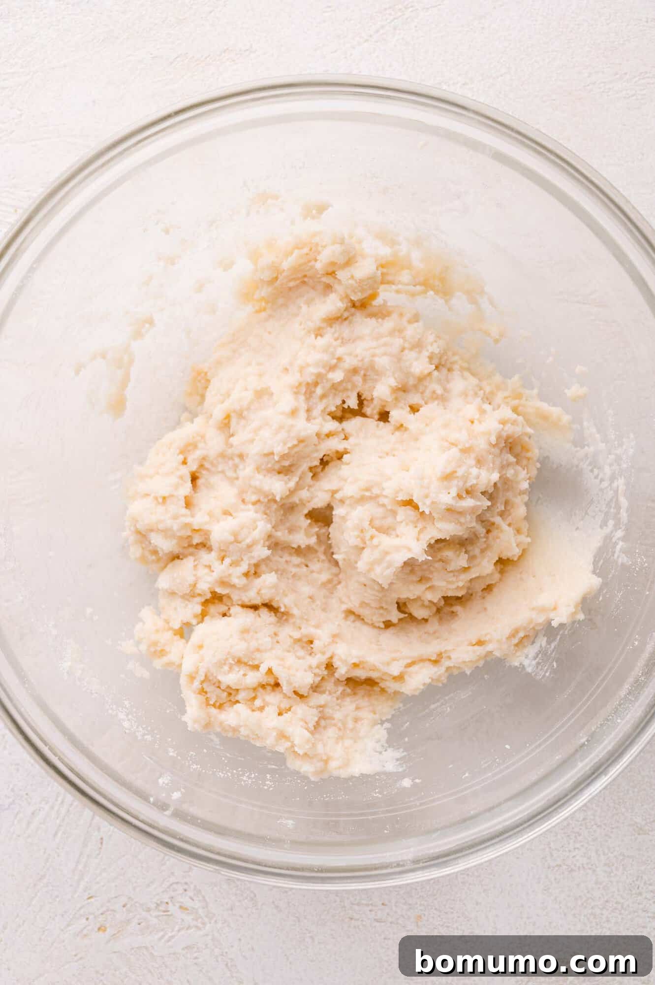 A clear glass bowl now holds a pale, thick, and cohesive dough mixture, exhibiting the sticky texture characteristic of a perfect cobbler topping, awaiting its final placement.