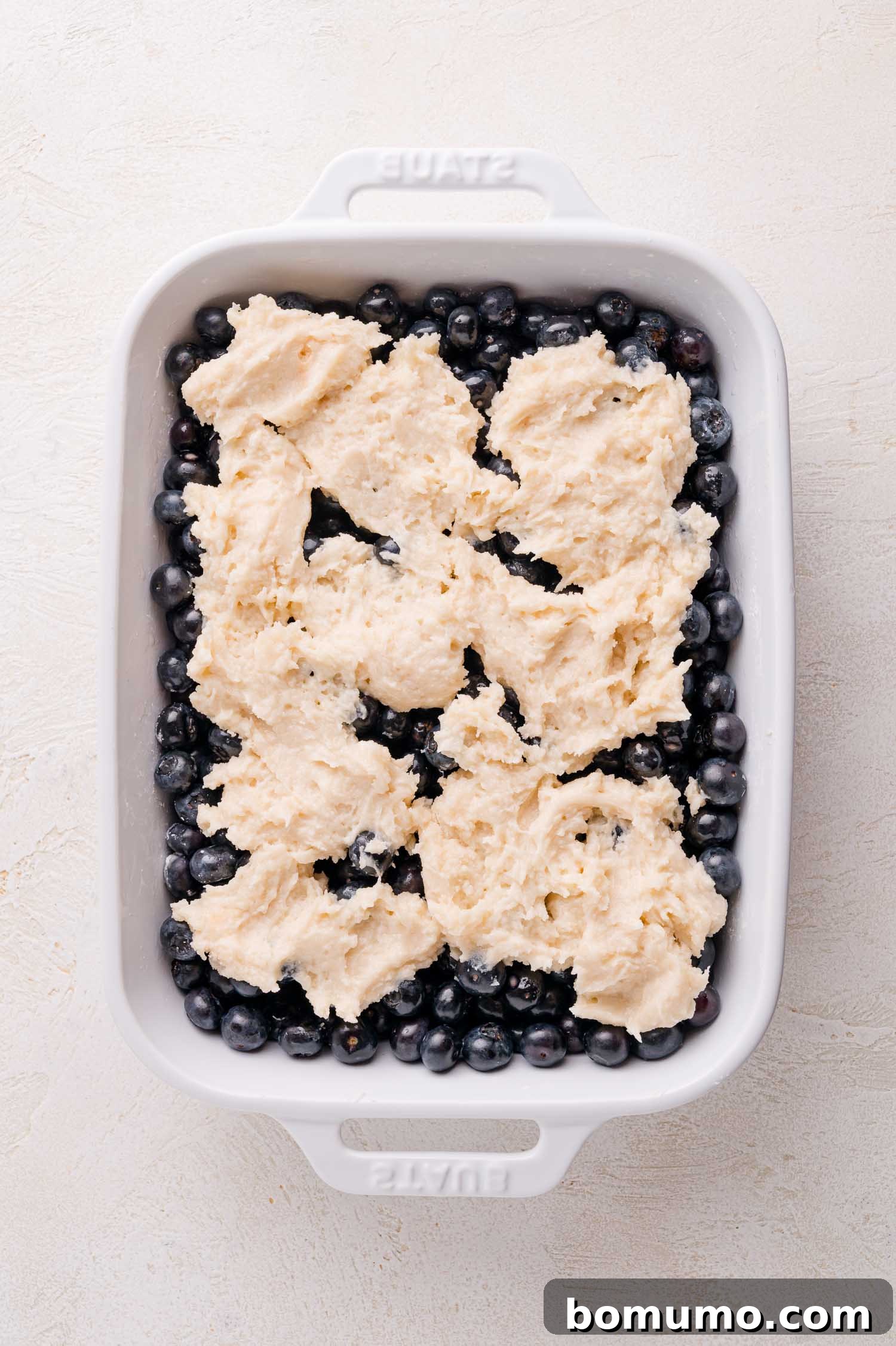 A white baking dish, now holding an artful arrangement of fresh blueberries beneath irregularly shaped dollops of biscuit dough, perfectly poised for baking.