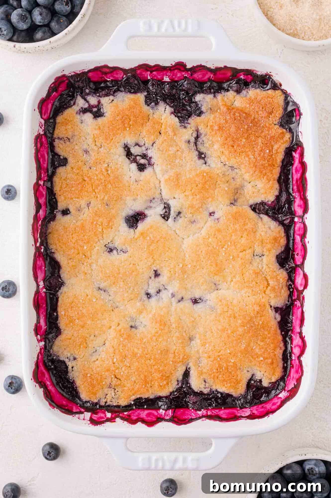 A beautifully baked blueberry cobbler, its golden-brown crust glistening in a pristine white rectangular dish, surrounded by a scatter of fresh blueberries and small, ingredient-filled bowls.